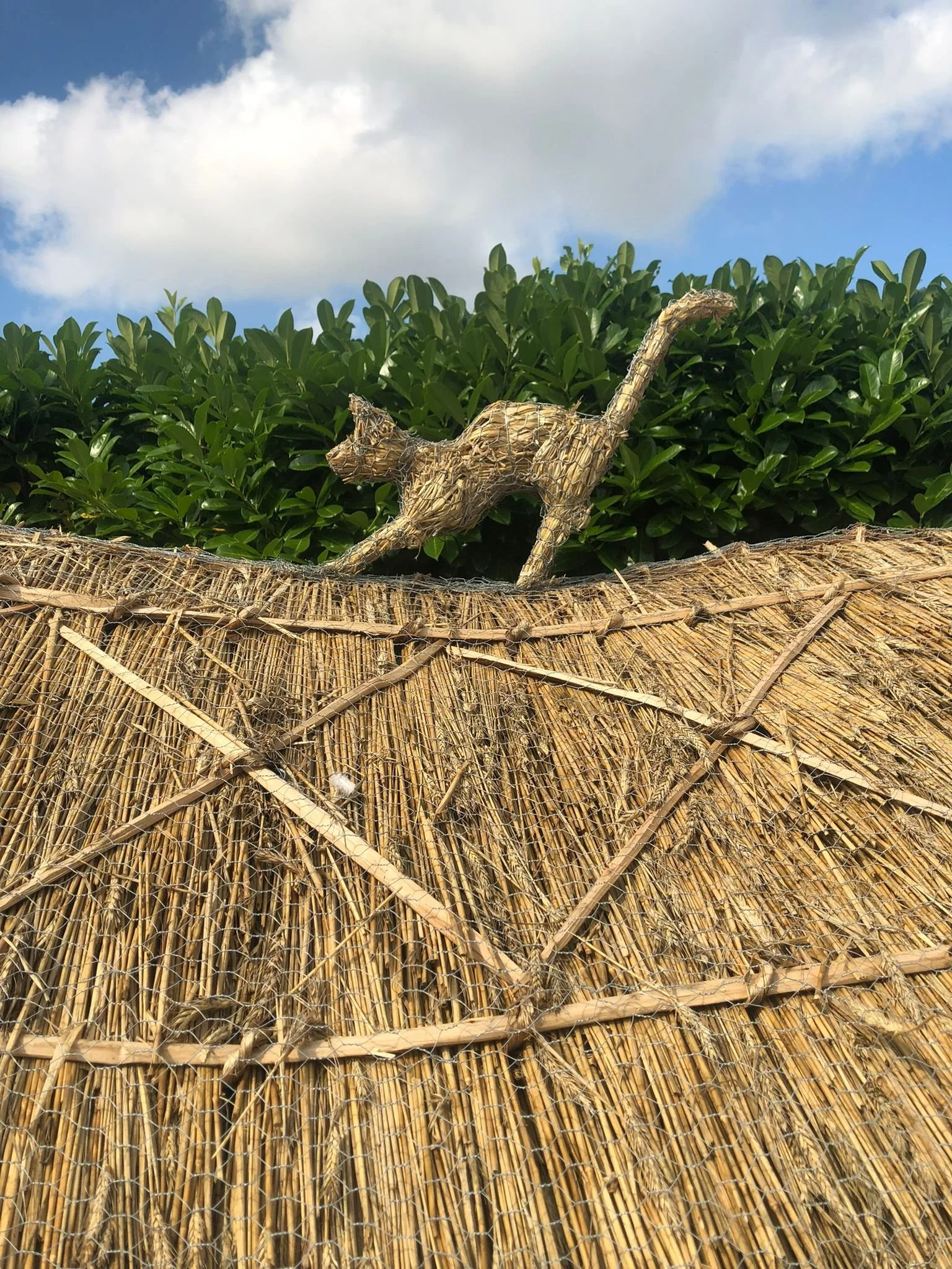 A woven straw cat sculpture perched on a thatched roof with a green hedge and a cloudy sky in the background.