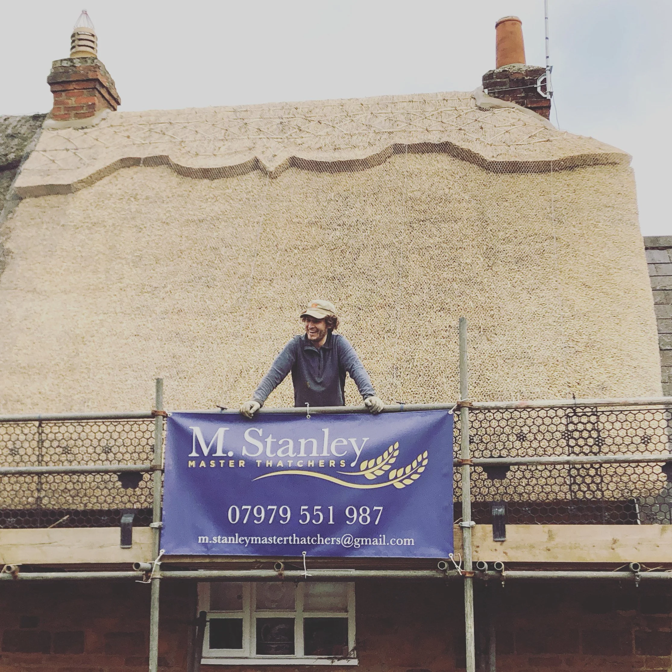 A person standing on scaffolding in front of a house wall under renovation, holding a sign for M. Stanley Master Thatchers, with contact information