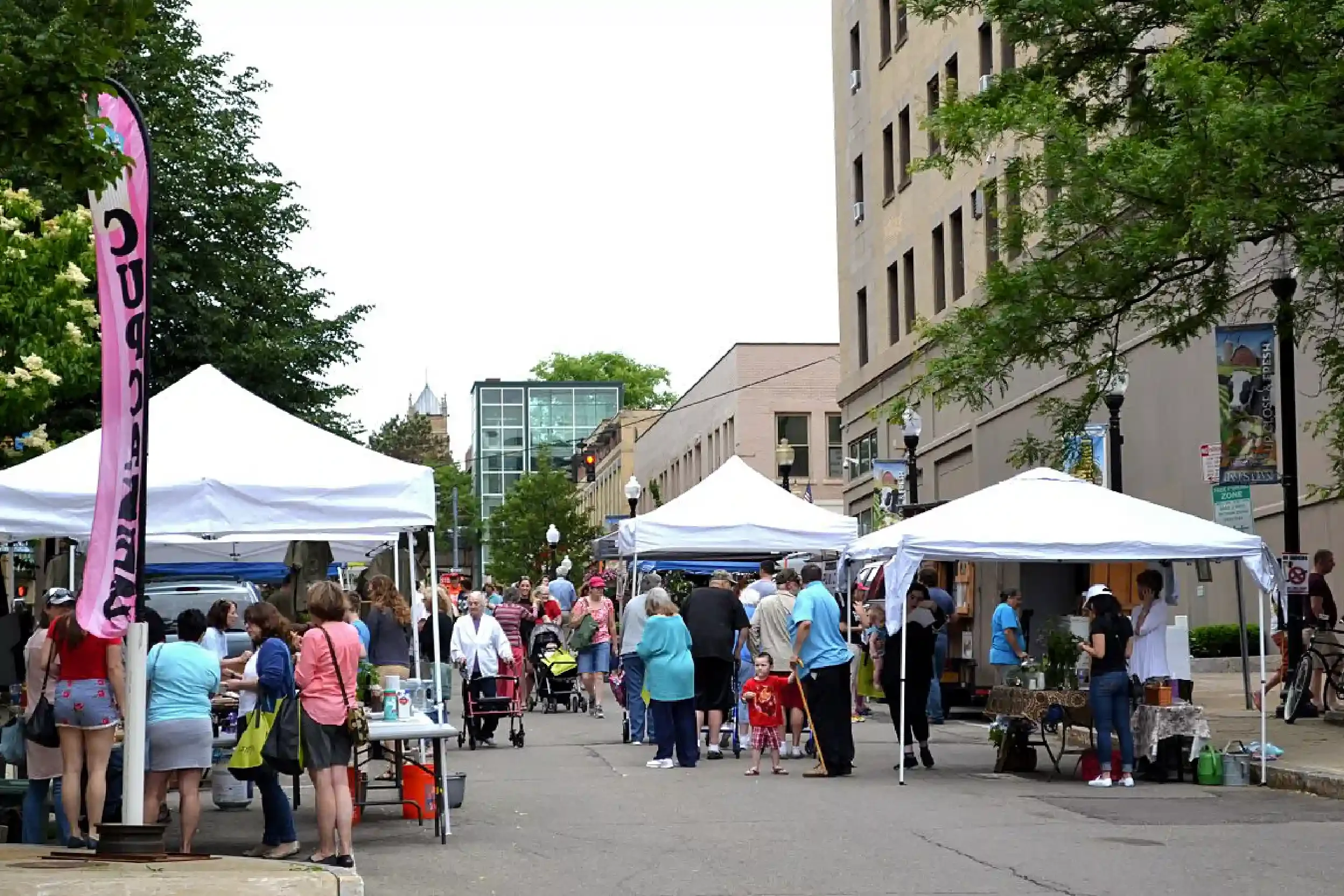 A busy outdoor market in a city street with white tents and many people walking. Tall buildings and trees are visible in the background.