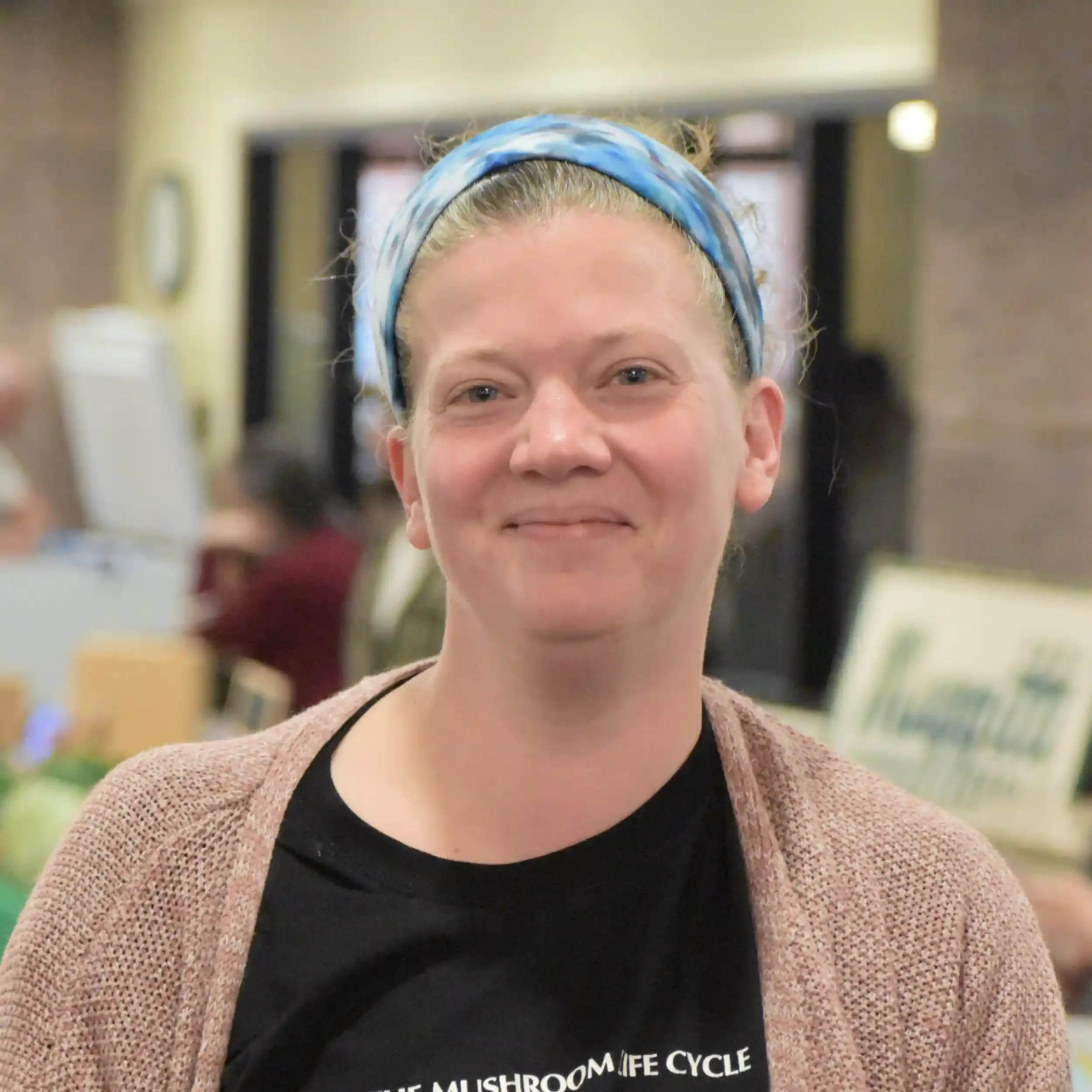 Close-up of a person's face, wearing a blue and white patterned headband, smiling, with a black T-shirt and a brown sweater, blurred indoor background.