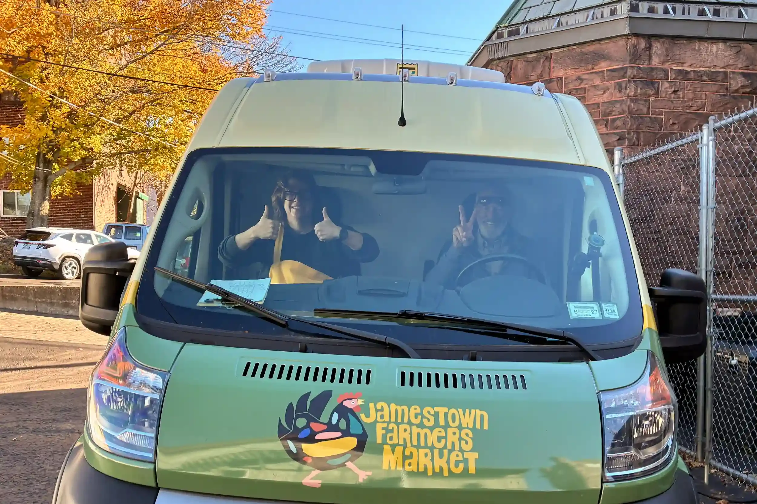 Front view of a green vehicle with a logo and text that reads "Jamestown Farmers Market" on the front. Two smiling people inside the vehicle are giving thumbs up and raising one finger. Background shows trees with autumn leaves, parked cars, brick building, and chain-link fence.