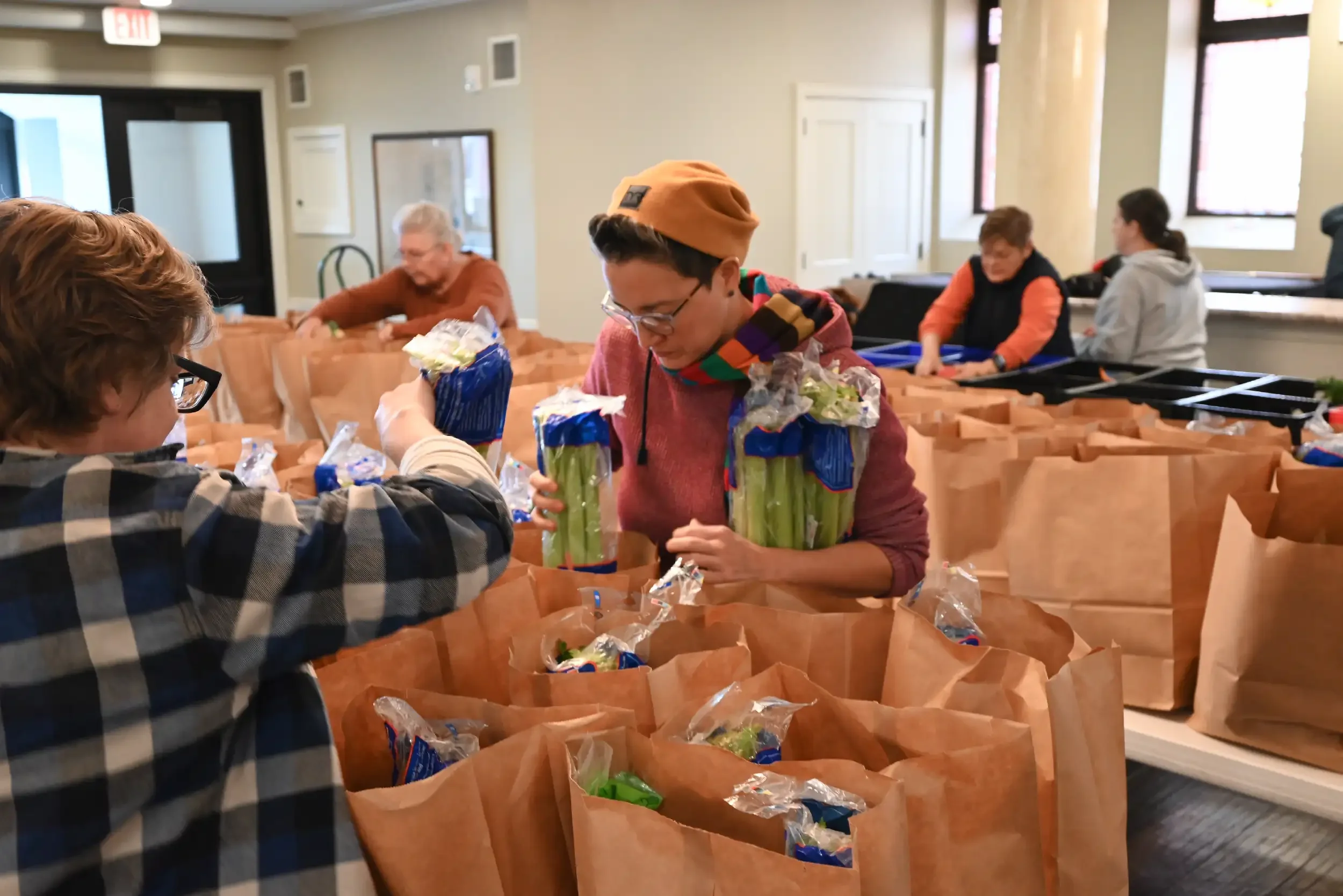 People packing groceries into paper bags in a community room with volunteers working at tables.