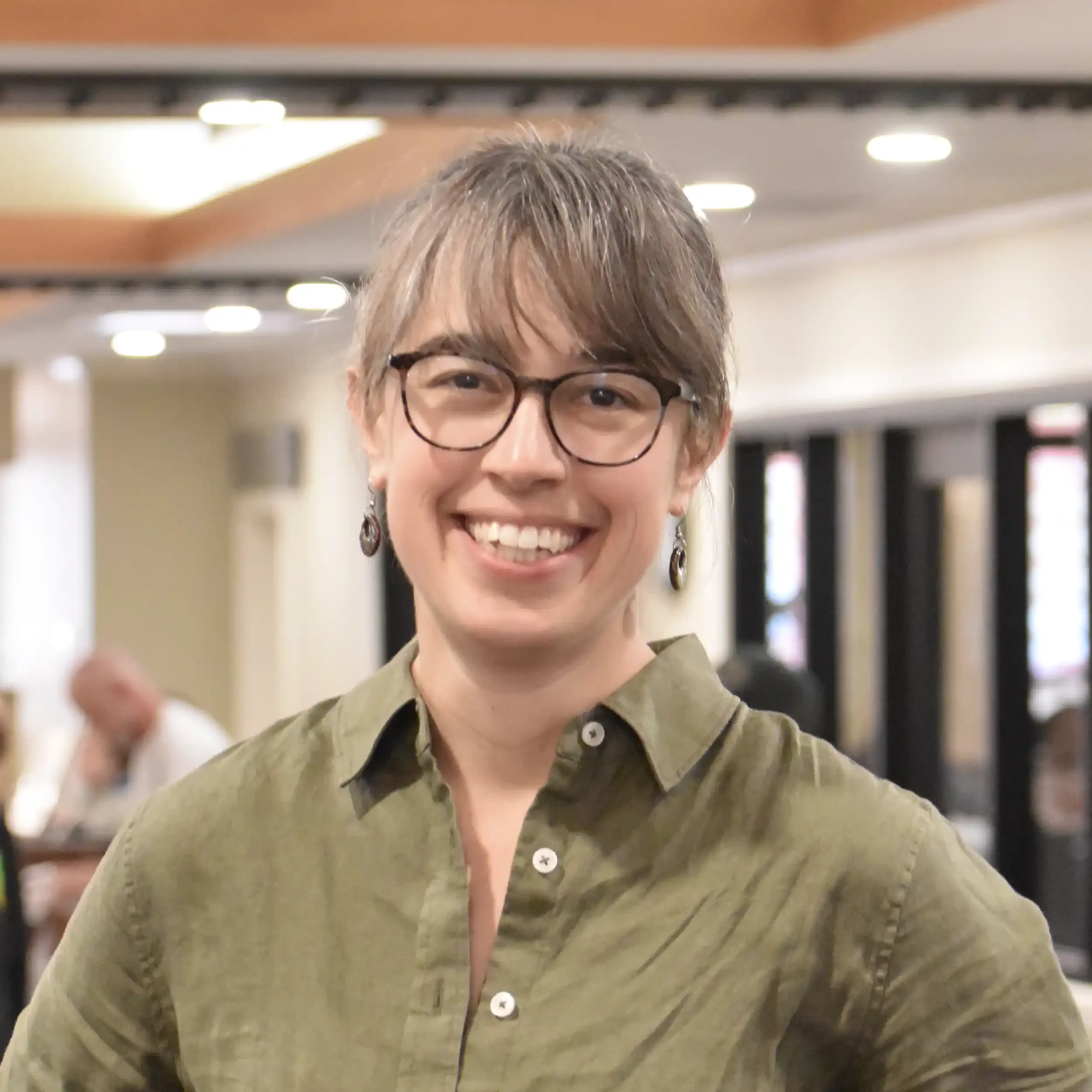 Close-up of a smiling person with short, light brown hair, wearing glasses, and an olive-green button-up shirt, in an indoor setting with blurred lights in the background.