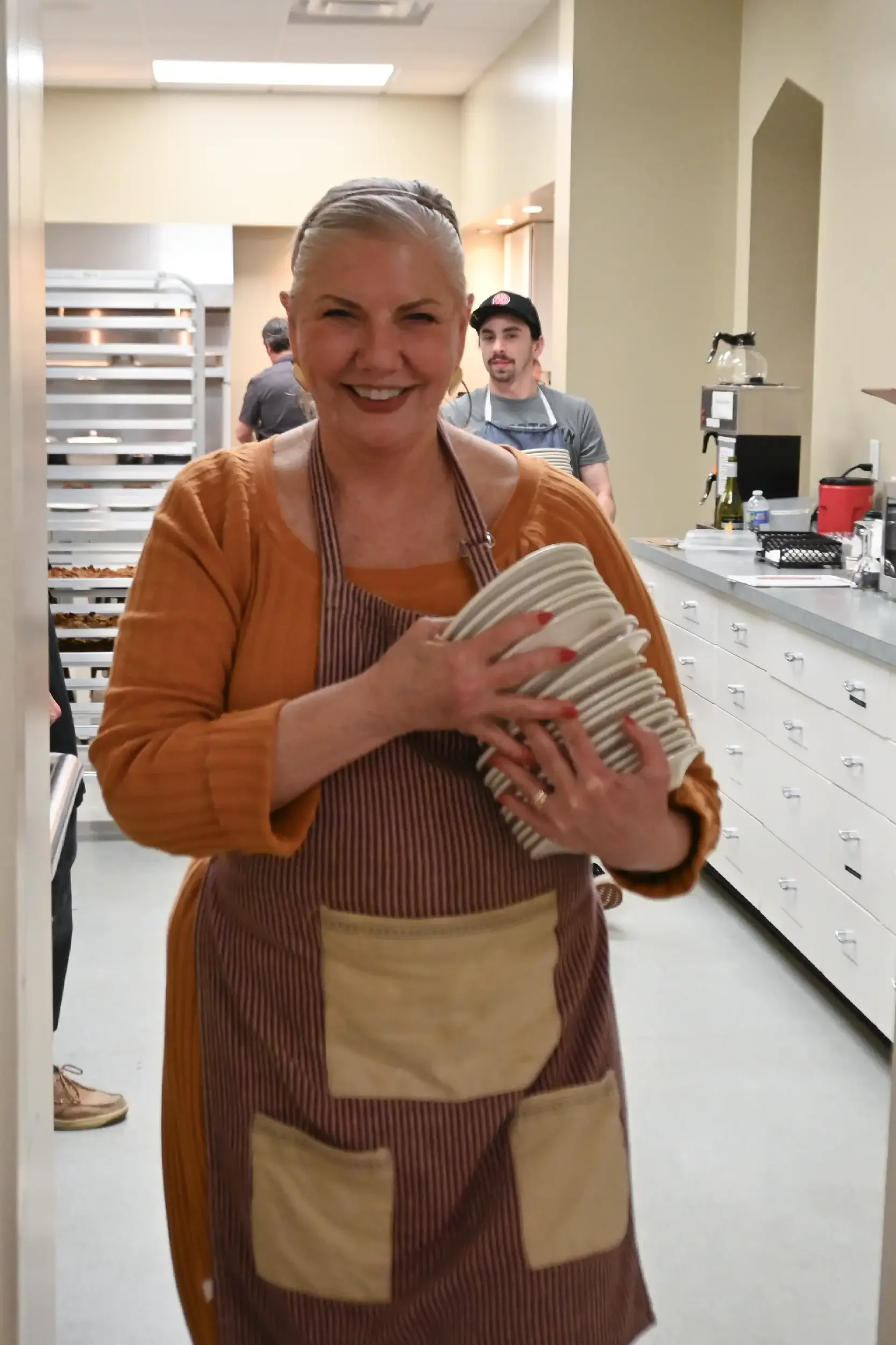 A smiling woman wearing an apron holds a stack of plates inside a kitchen or bakery. Two men are visible in the background, one wearing a hat and apron, busy in the kitchen area.
