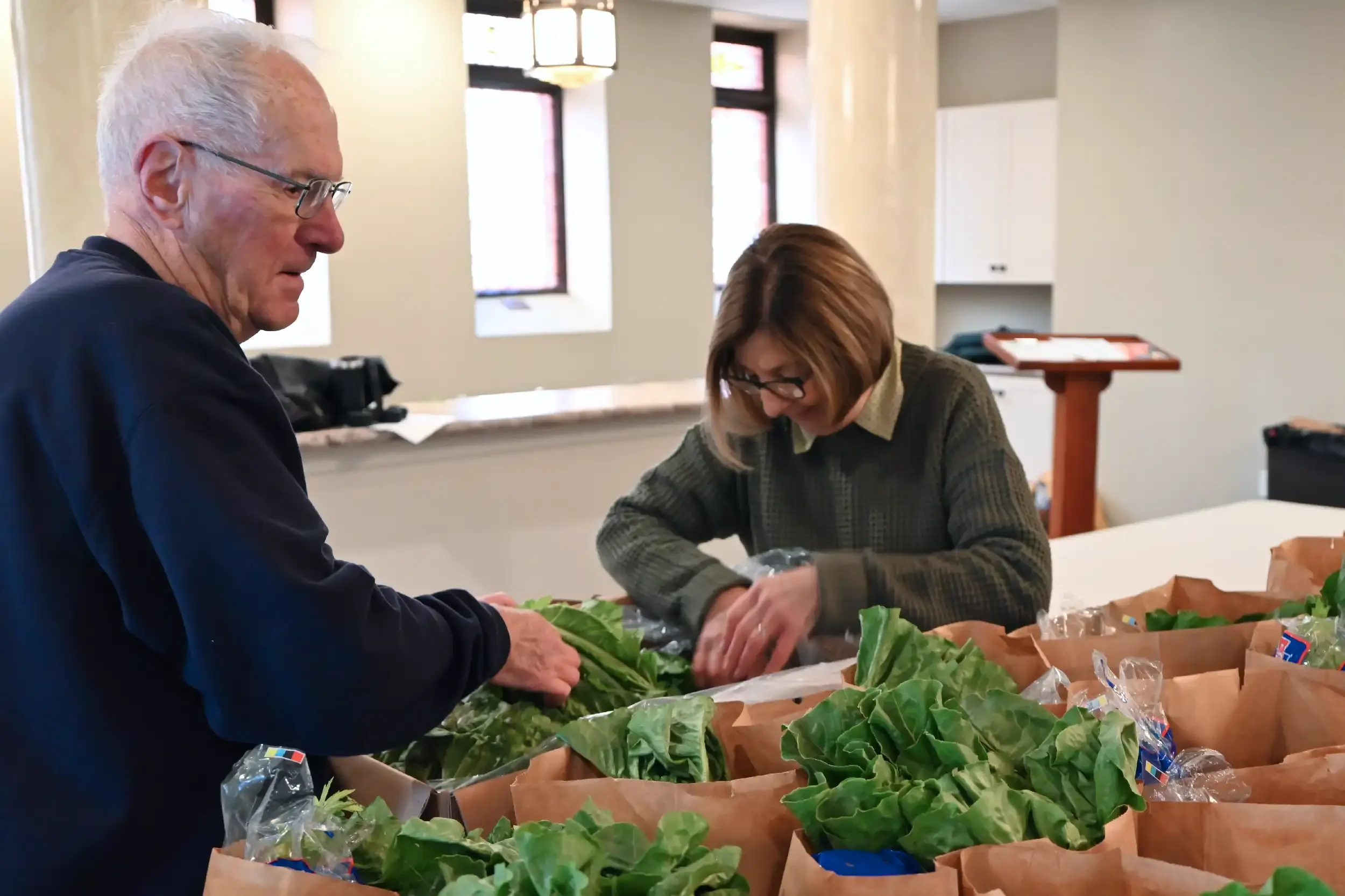 Two older adults, a man with white hair and a woman with brown hair, both wearing glasses, are working with leafy greens on a table filled with grocery bags in an indoor market or store.