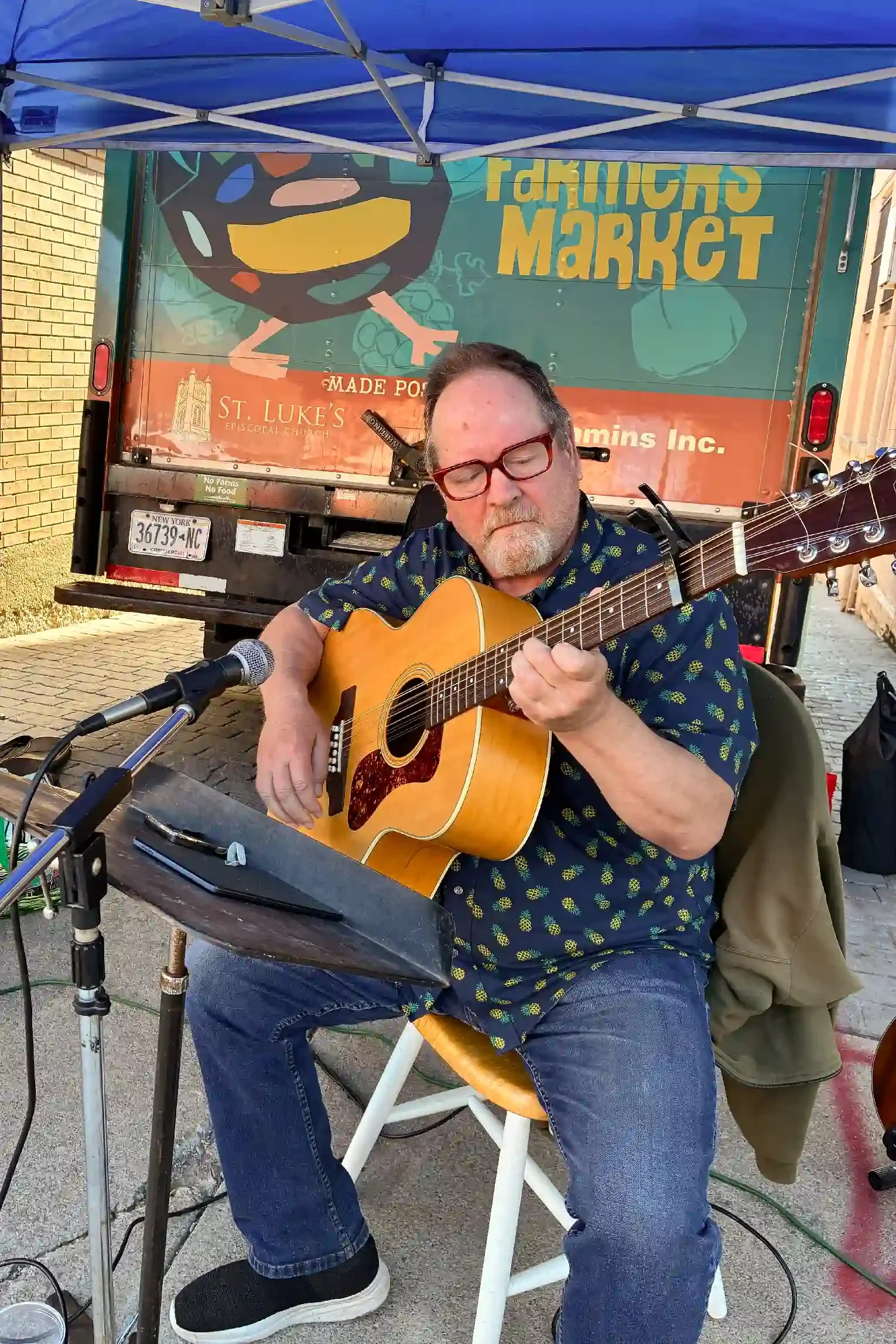 A man with glasses playing an acoustic guitar and singing into a microphone at an outdoor market. Behind him is a truck with a colorful sign that reads 'Farmer's Market' and features a smiling face made of fruit and vegetables.
