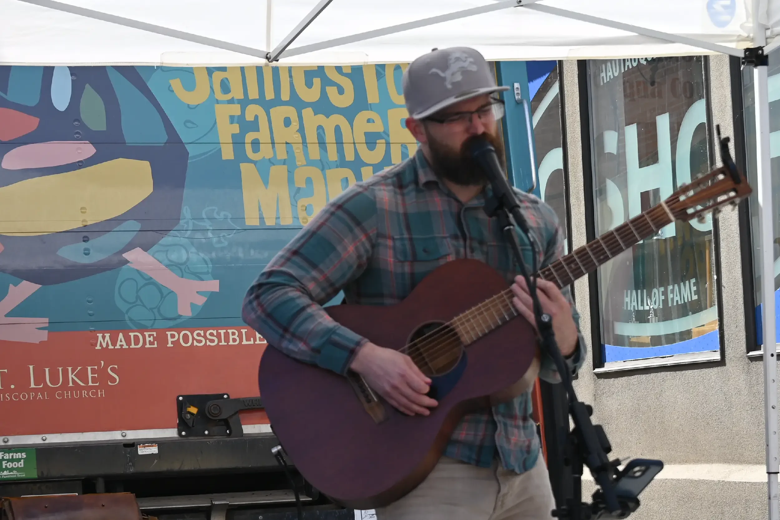A man wearing a gray cap, glasses, and a plaid shirt playing an acoustic guitar and singing into a microphone at an outdoor event, with a colorful banner and windows behind him.