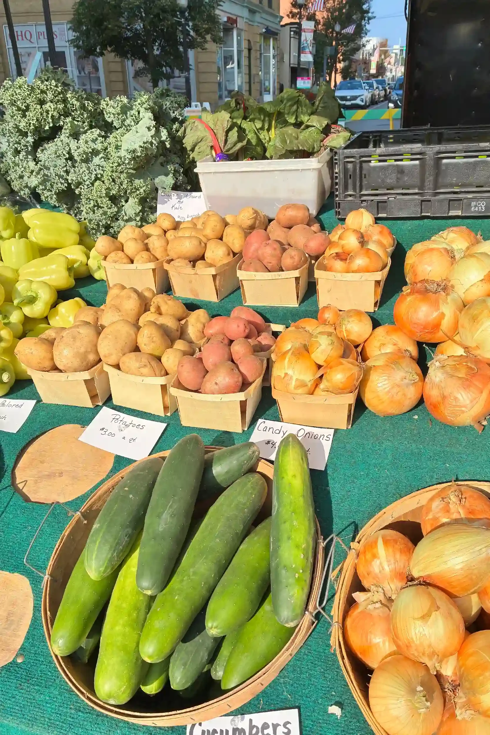 Fresh cucumbers in a basket, yellow bell peppers, potatoes, sweet potatoes, and onions displayed at an outdoor market stall with a city street in the background.