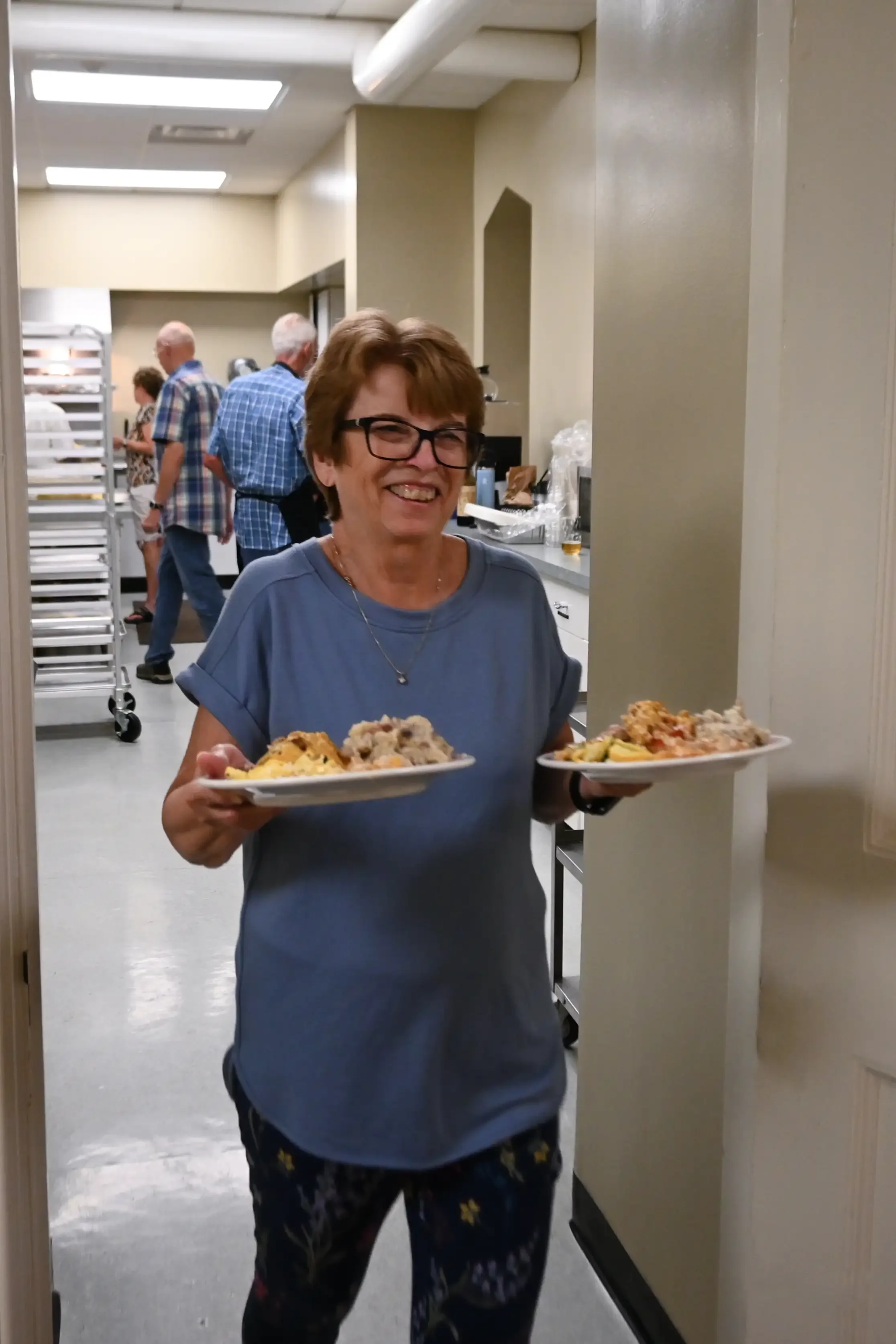 A woman smiling and holding two plates of food, walking through a kitchen or cafeteria with other people in the background.
