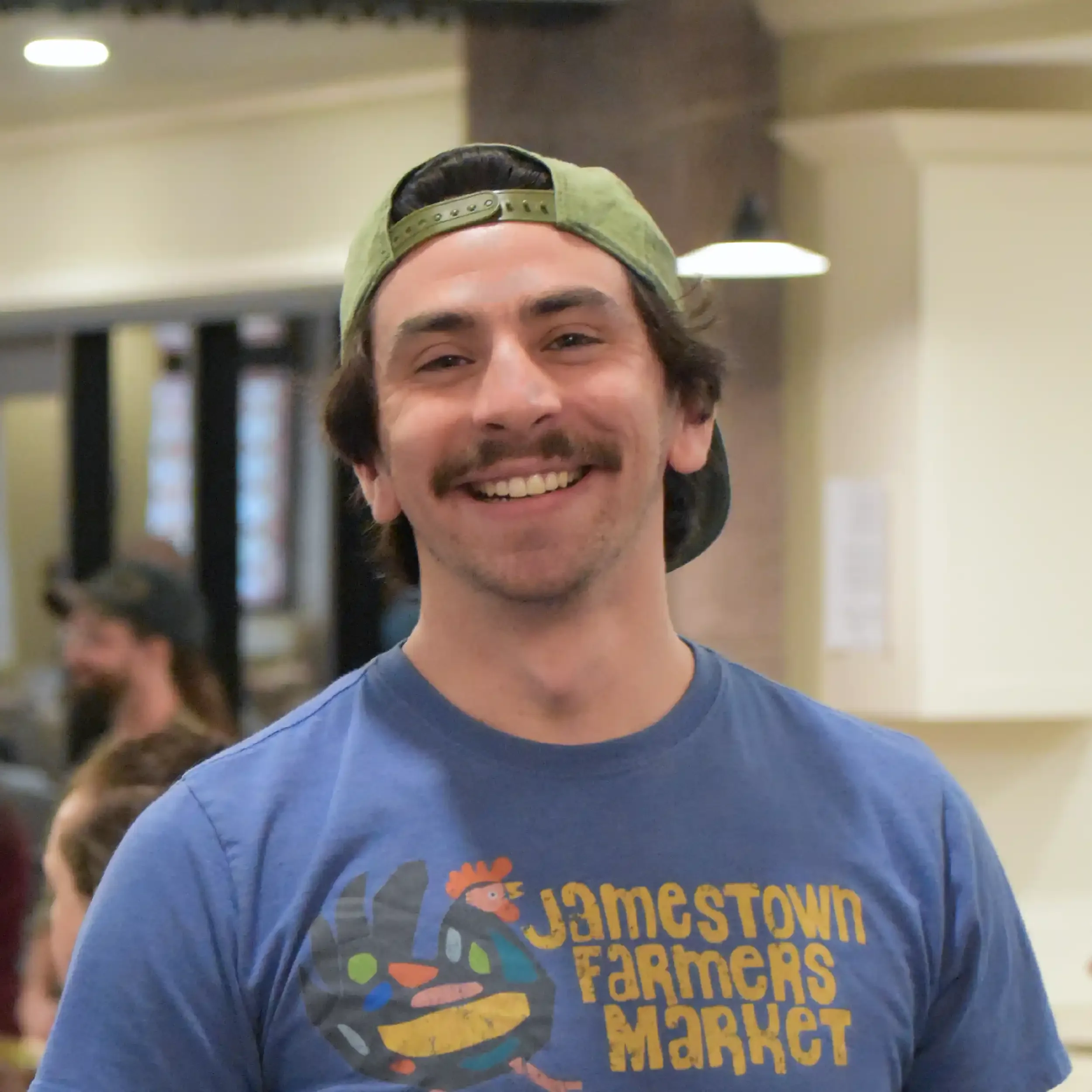 Smiling man wearing a green cap backwards and a blue T-shirt with colorful text that says 'James Town Farmers Market' in an indoor setting.
