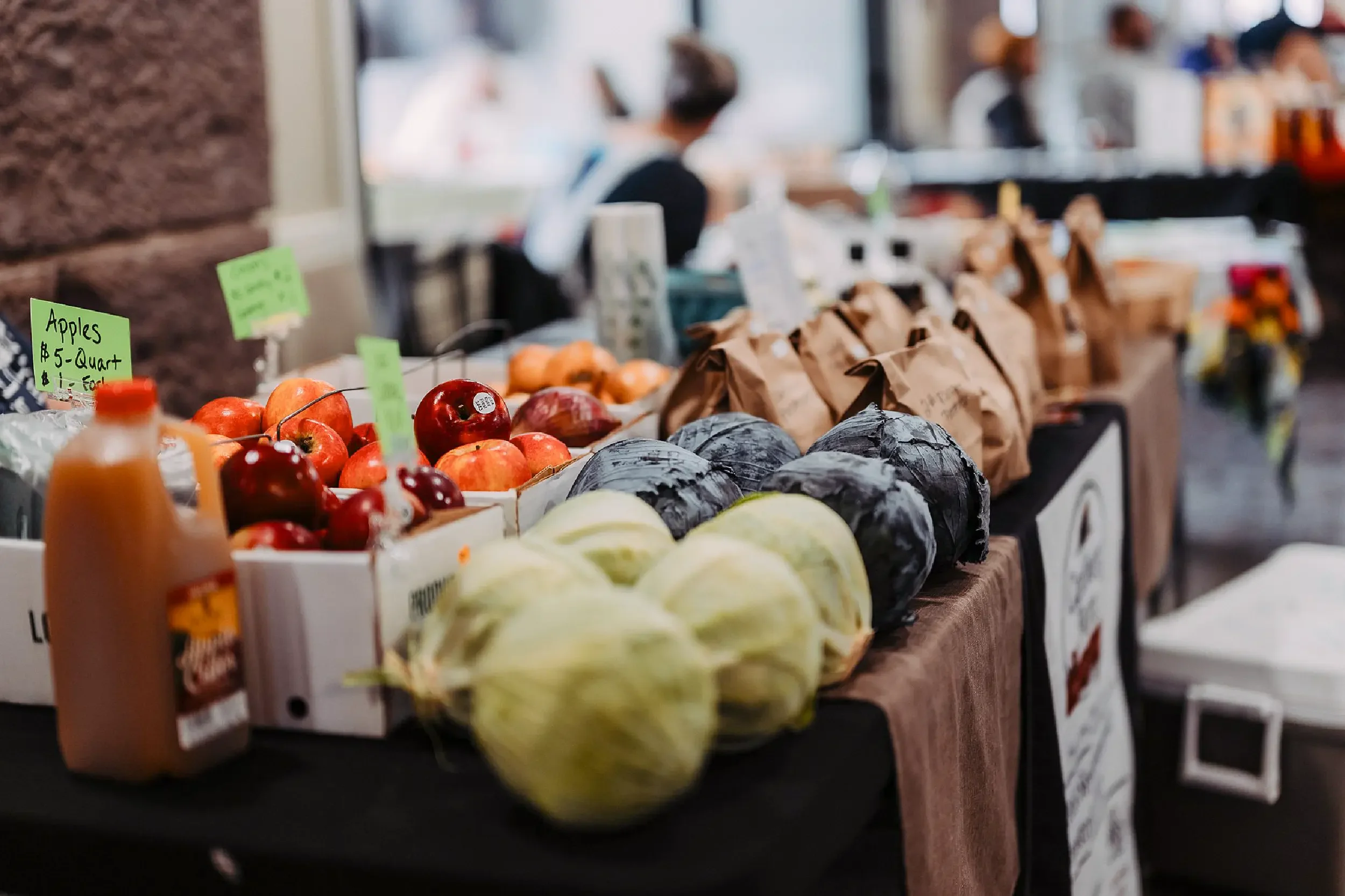 Produce stand with apples, cabbages, and handwritten price signs at an indoor farmers market.