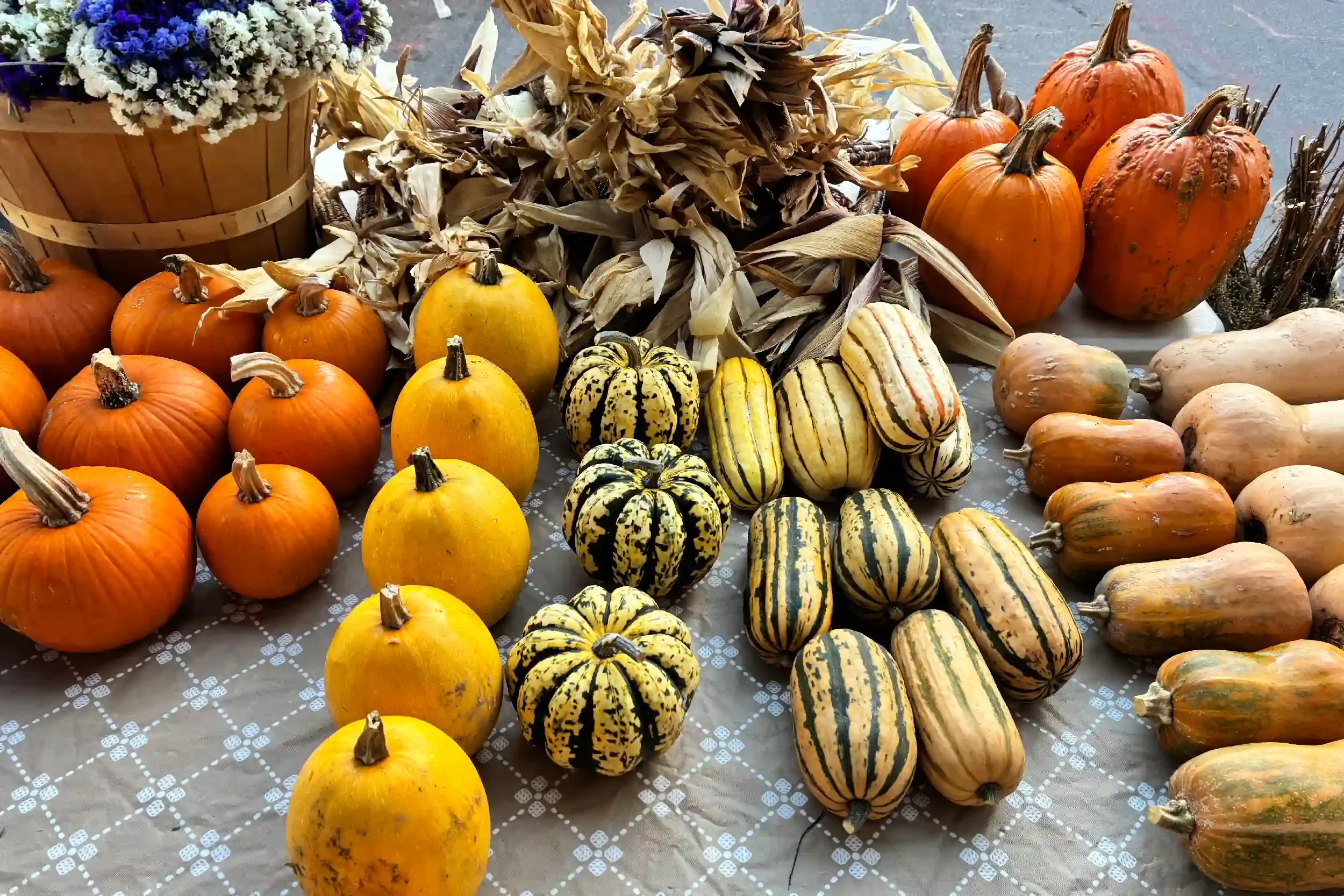 Fall display of pumpkins, gourds, dried corn, and dried leaves on a table, with a basket of white and purple flowers to the left.
