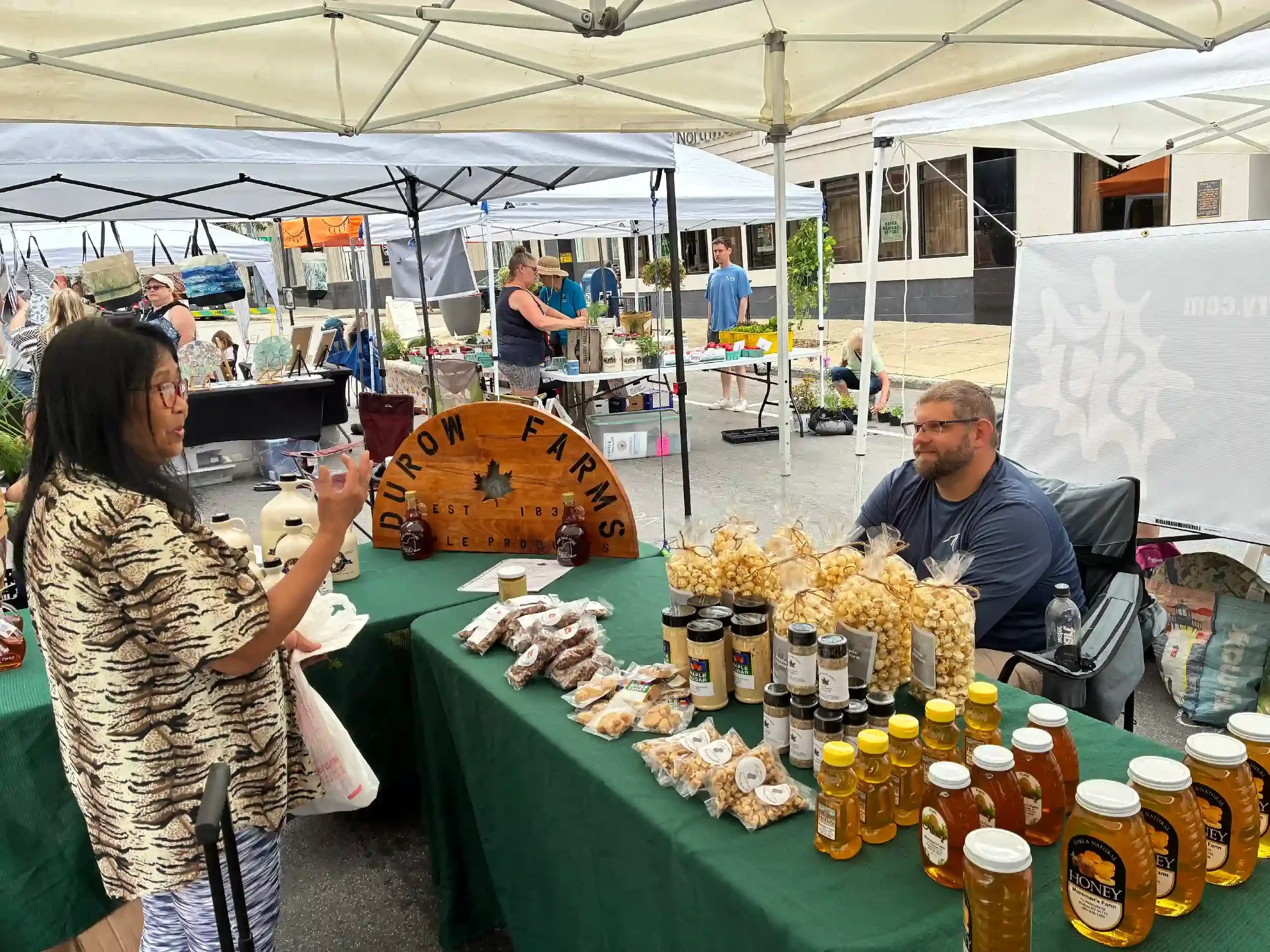 Scene from a street market with white tents and tables displaying honey jars, honey sticks, and packaged snacks. Several people browse and interact at the booths, with a woman in a tiger-print shirt and a man with glasses seated at a table in the foreground. The background shows other stalls, hanging fabrics, and a building with display windows.