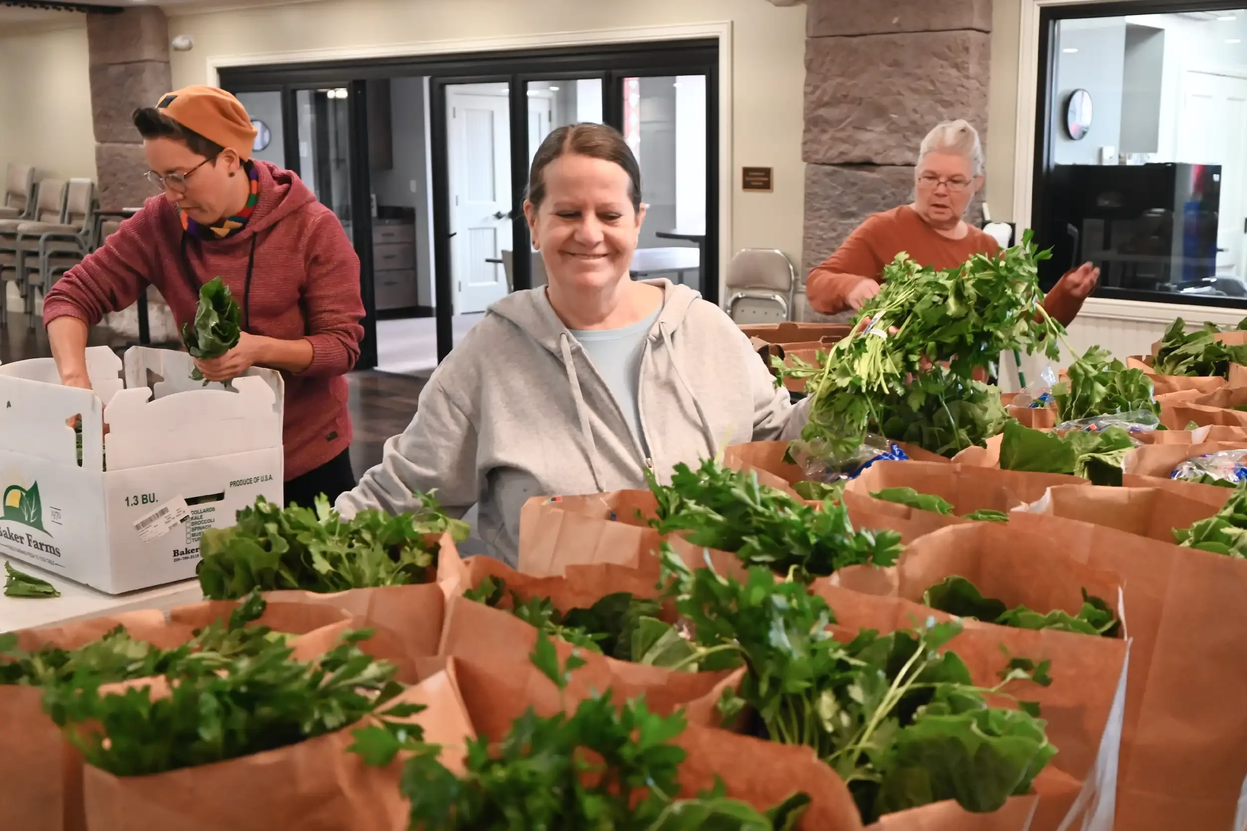 Three women in an indoor space sorting and packing fresh greens into paper bags, with white boxes in the foreground.