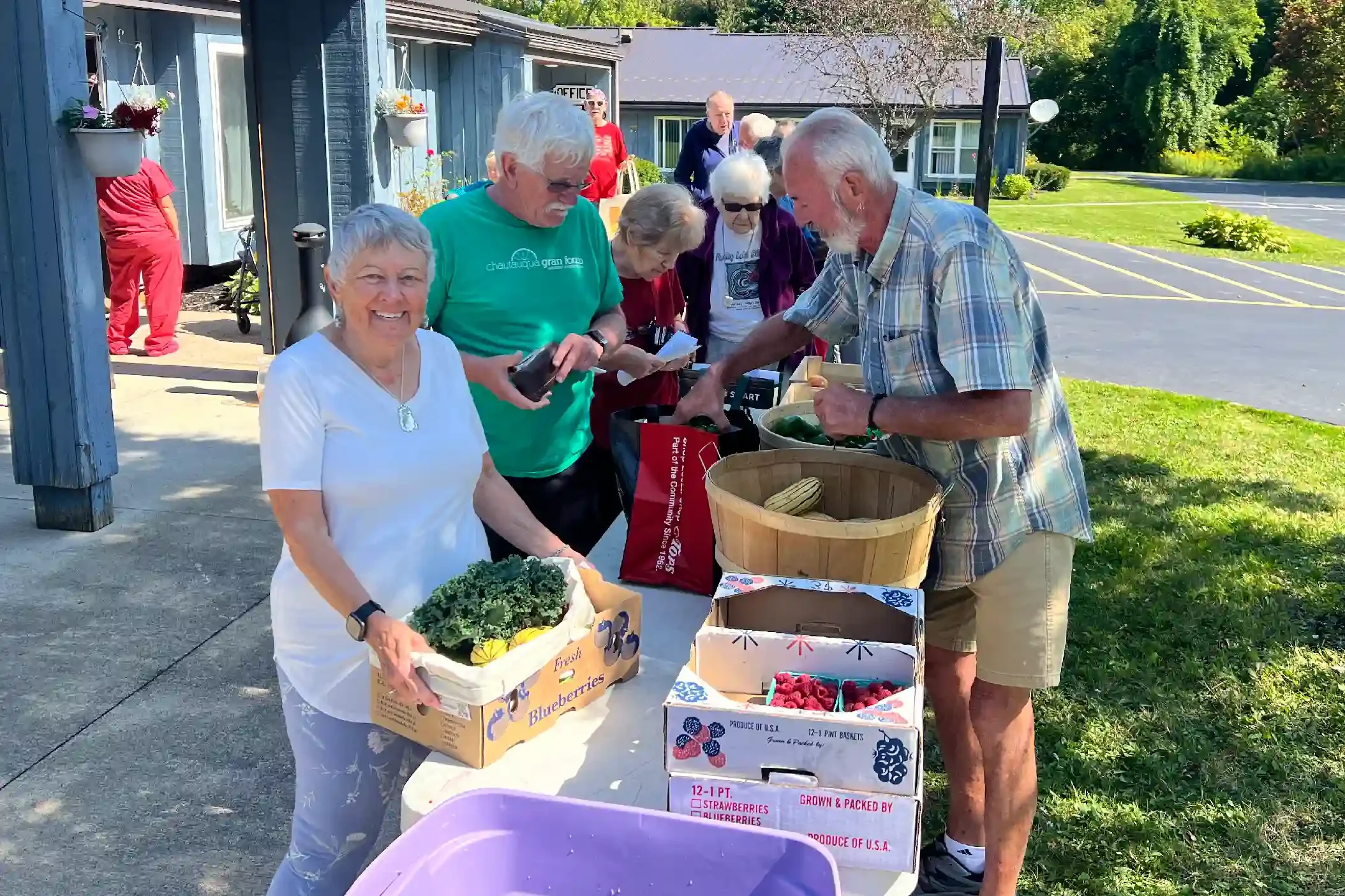 A group of people gathered outdoors at a farmers market or similar event, selling and buying fresh berries and produce. Some are smiling and browsing the items. There are boxes of blueberries, strawberries, and raspberries on the table.