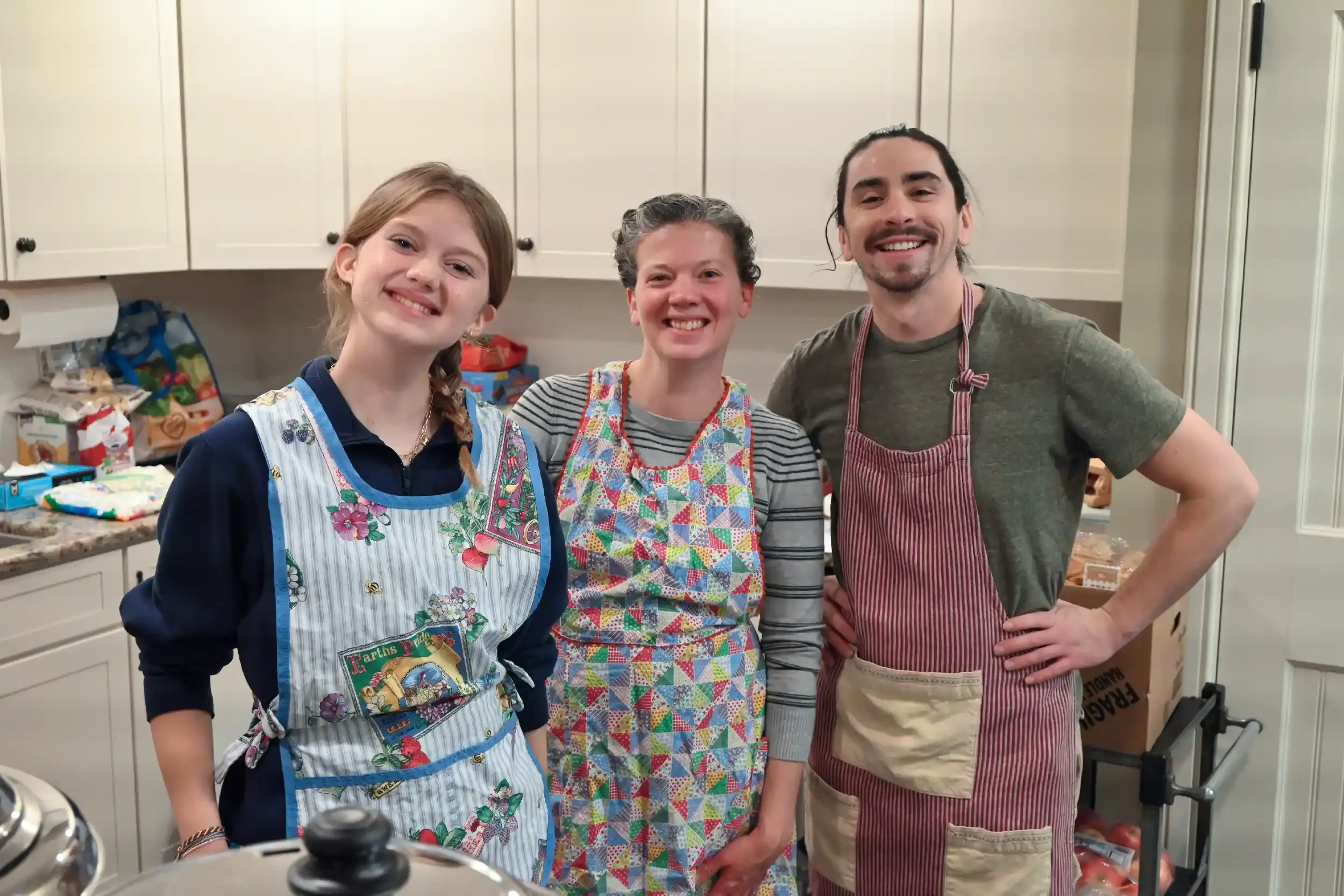 Three people smiling and wearing aprons in a kitchen, standing close together with their arms around each other.