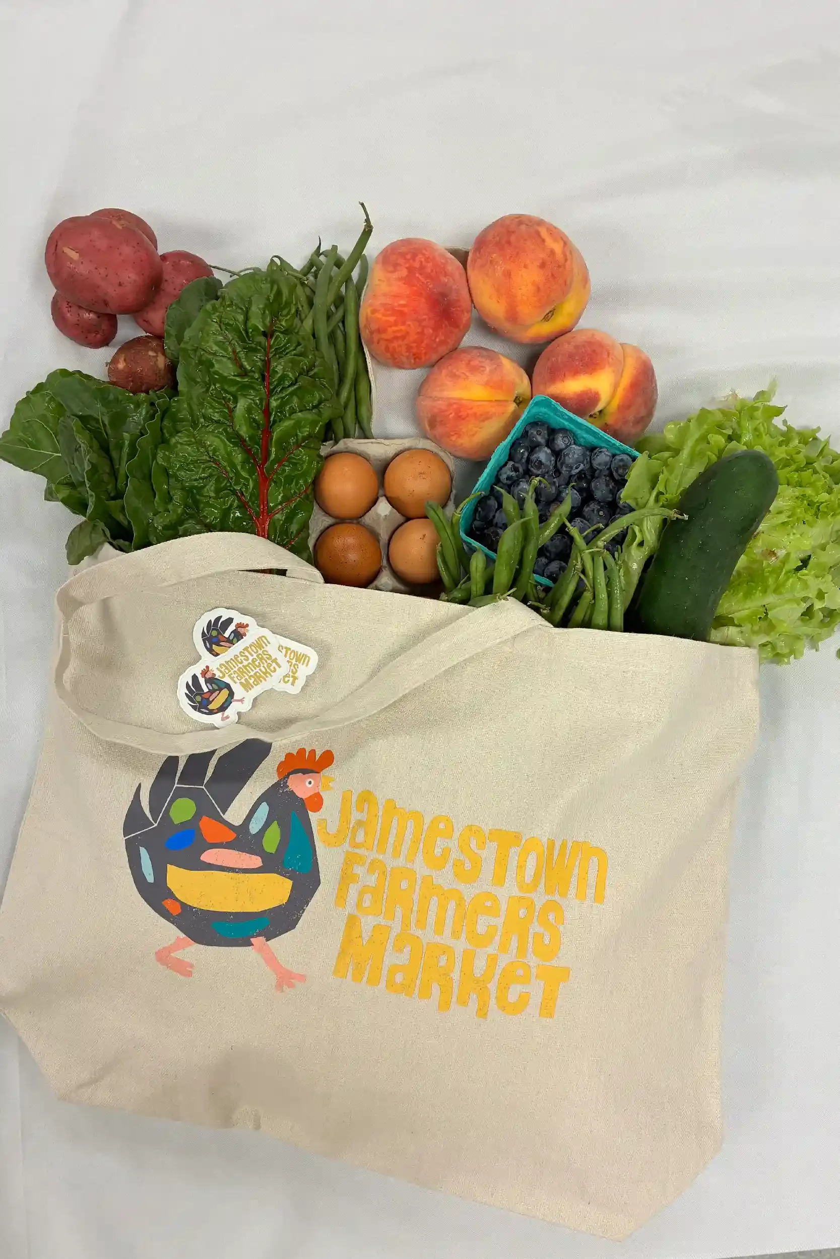 A tote bag with a colorful chicken illustration and the text 'Jamestown Farmers Market' printed on it, filled with fresh fruits and vegetables including peaches, blueberries, green beans, cucumbers, potatoes, and leafy greens.