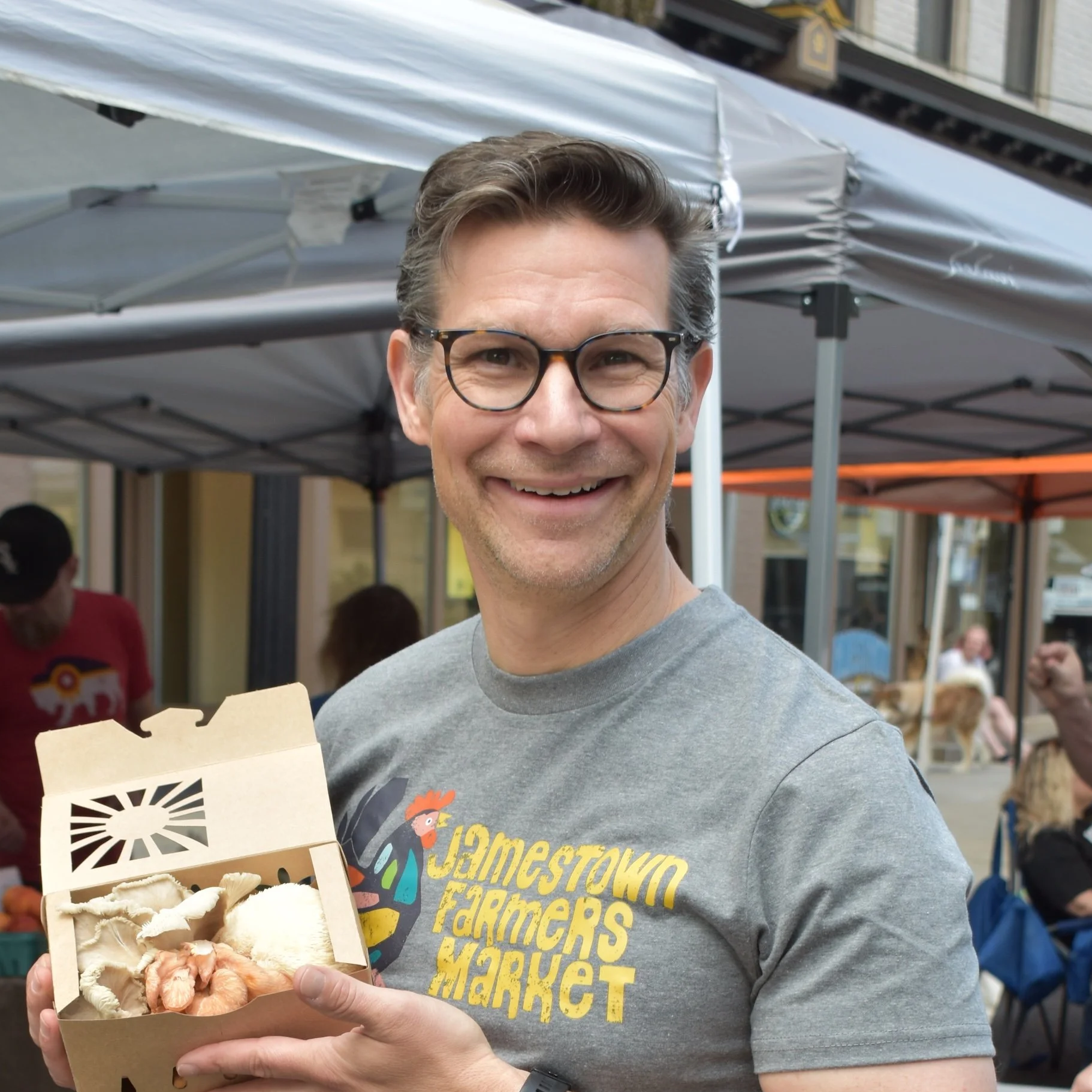 Close-up of a smiling man wearing black-rimmed glasses, with brown hair and a face full of wrinkles, dressed in a light blue zip-up sweater over a white shirt.