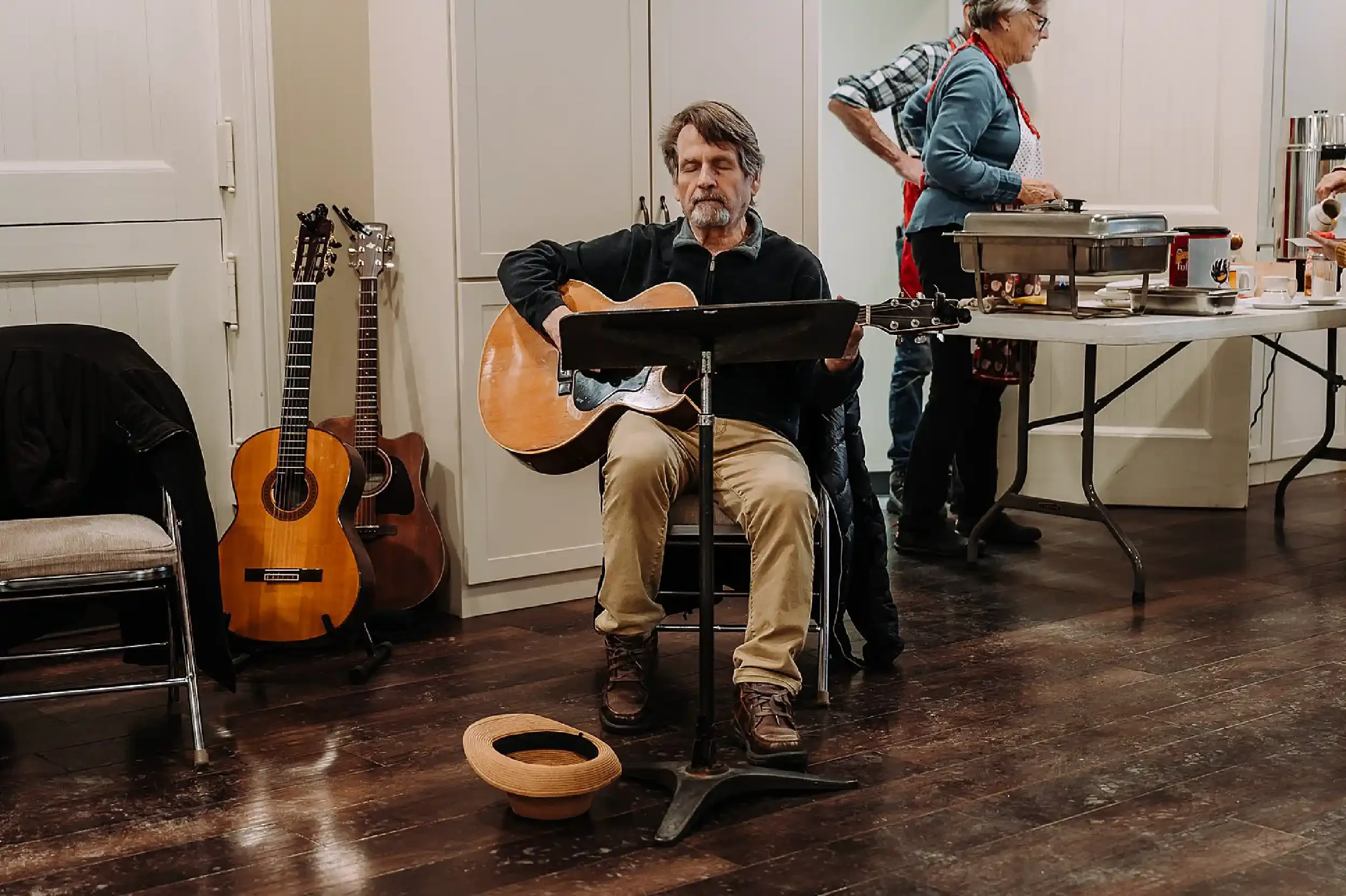 An older man playing an acoustic guitar while seated, with two additional guitars standing nearby, in a kitchen setting. In the background, a woman is preparing food at a table, and part of another person is visible on the right.