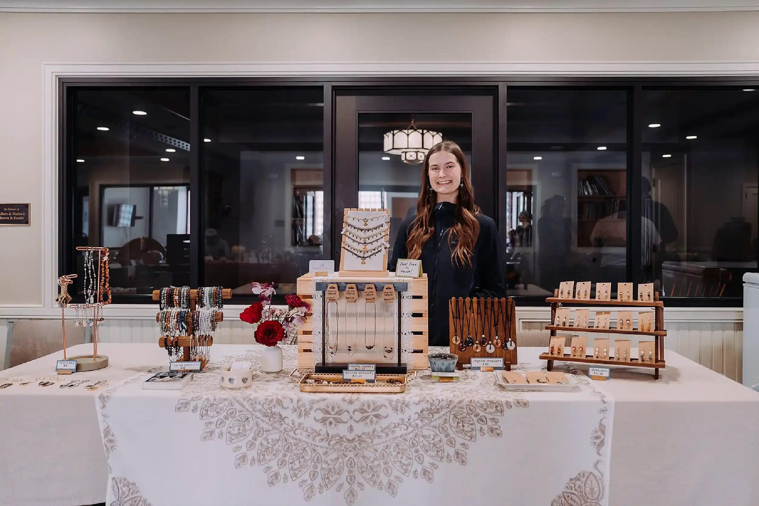A woman stands behind a jewelry table at an indoor marketplace. The table displays necklaces, bracelets, and earrings with small signs and prices, and a small flower arrangement is on the table. Behind her is a large black-framed glass window with the interior of a building visible.