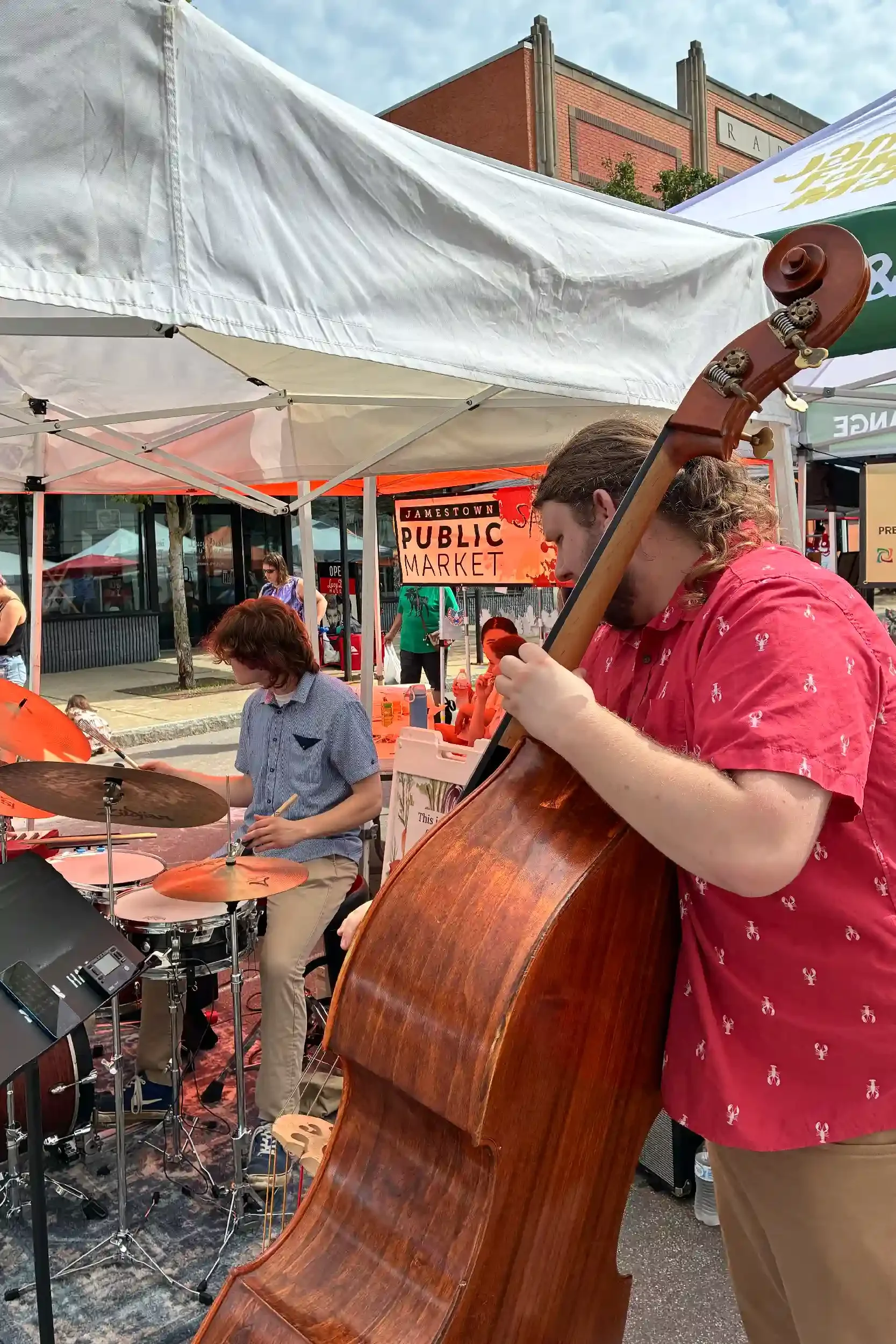 Two musicians performing at a farmers market, with a drummer and a bassist on stage under white tents.