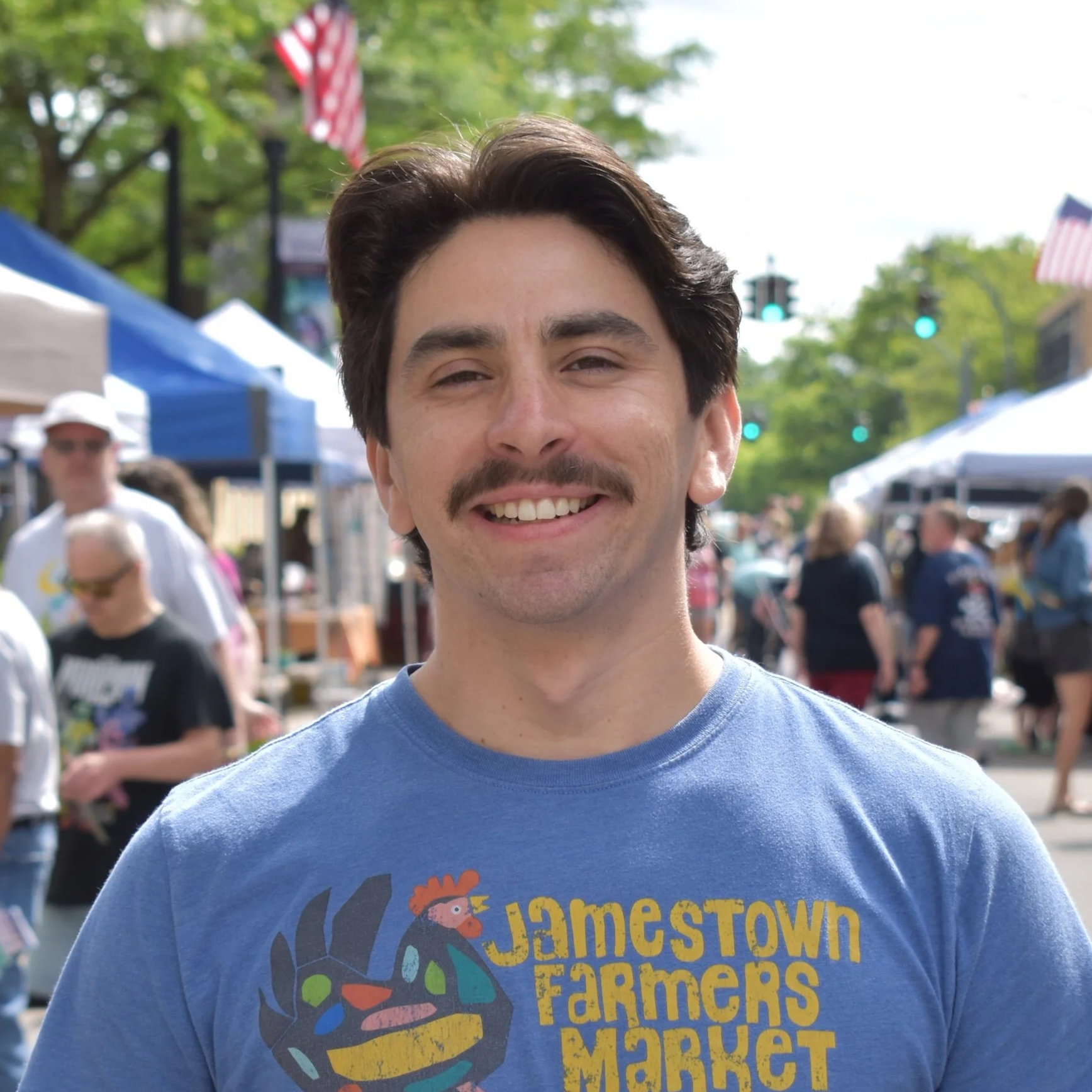 Smiling man wearing a green cap backwards and a blue T-shirt with colorful text that says 'James Town Farmers Market' in an indoor setting.