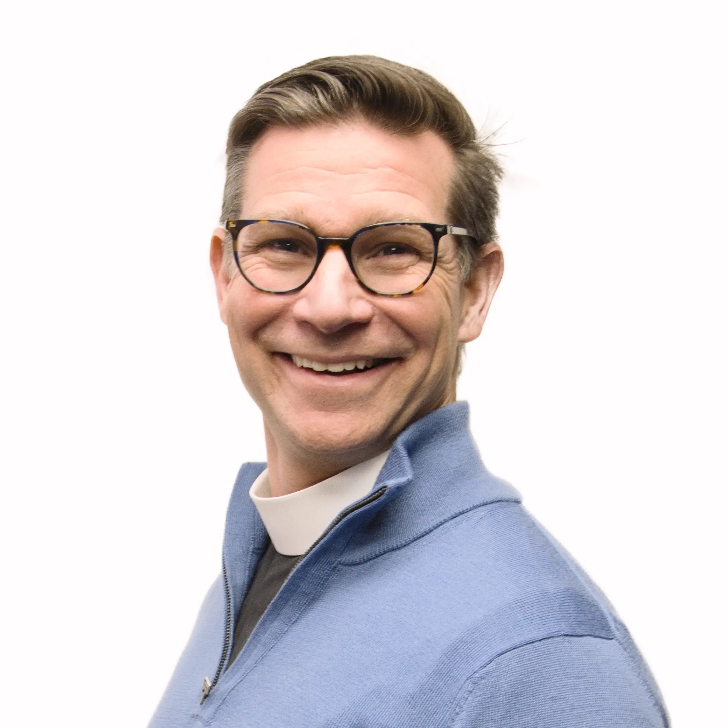 Close-up of a smiling man wearing black-rimmed glasses, with brown hair and a face full of wrinkles, dressed in a light blue zip-up sweater over a white shirt.