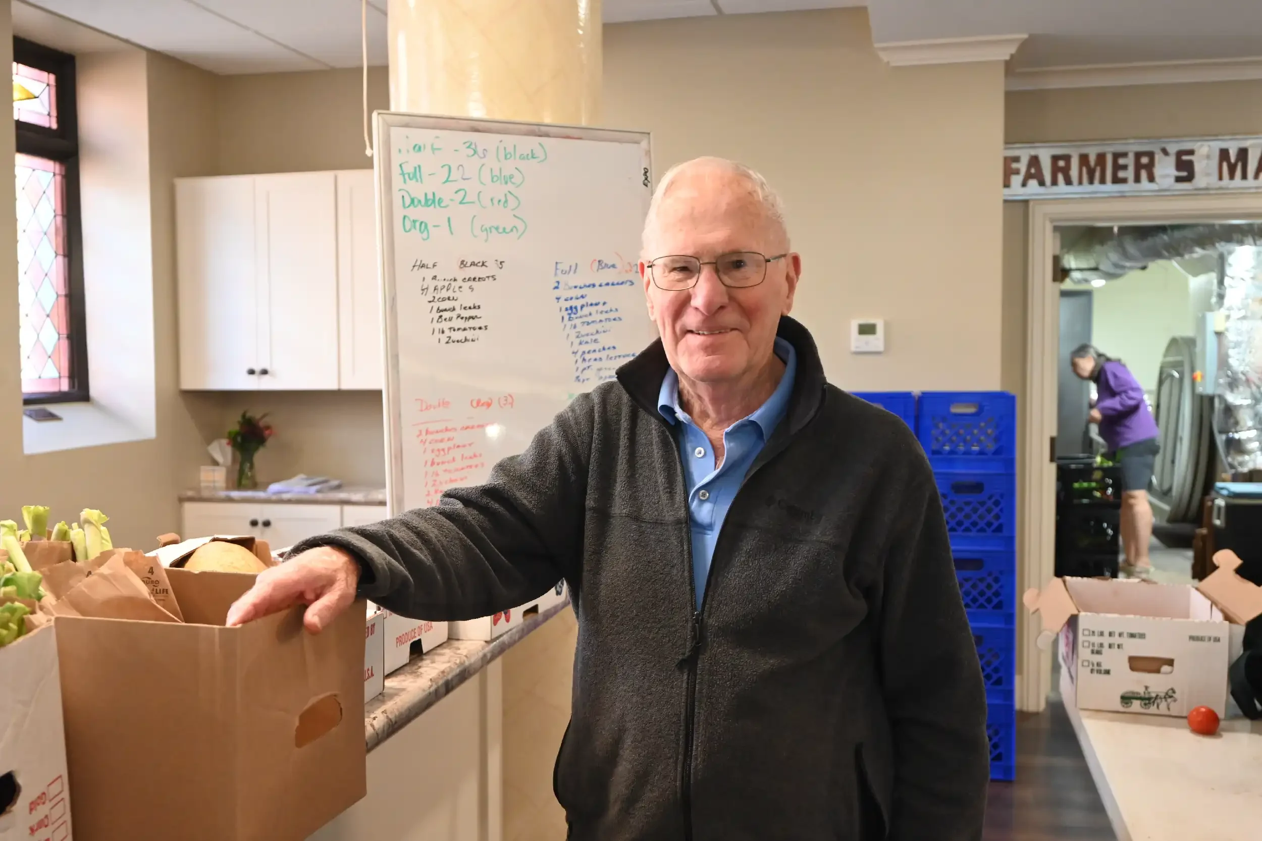 An elderly man with glasses and a gray sweater smiling at the camera while standing at a counter with brown paper bags filled with produce, in a room with white cabinets and a whiteboard with writing on it, and a woman in a purple jacket working in t