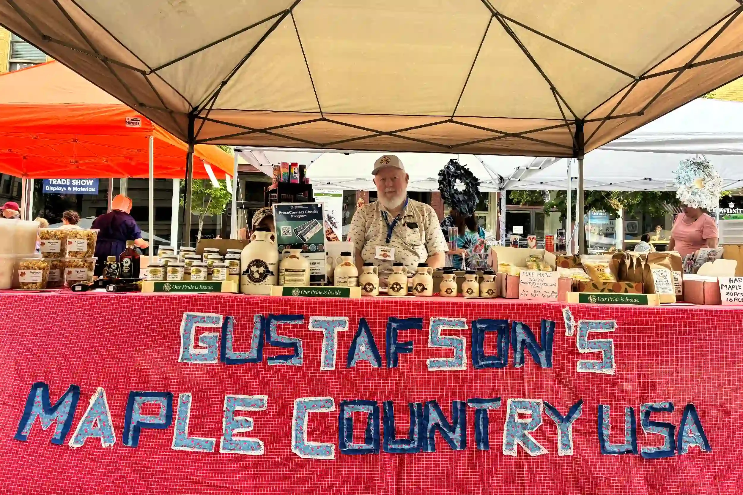 A vendor stands behind a table at an outdoor market stall selling honey and maple syrup products, with colorful signs and a red tablecloth displaying the products.