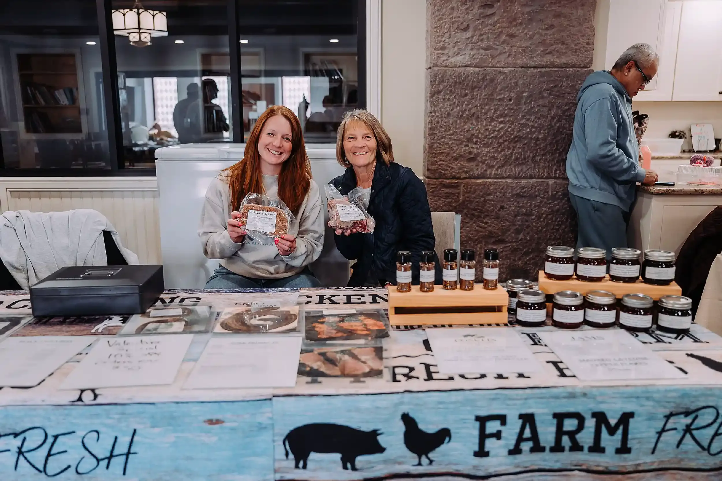 Two women sitting behind a table at a farmers market, holding packaged meats and smiling. The table displays jars, brochures, and a sign with farm-related words. A man is in the background, looking at products in a kitchen area.