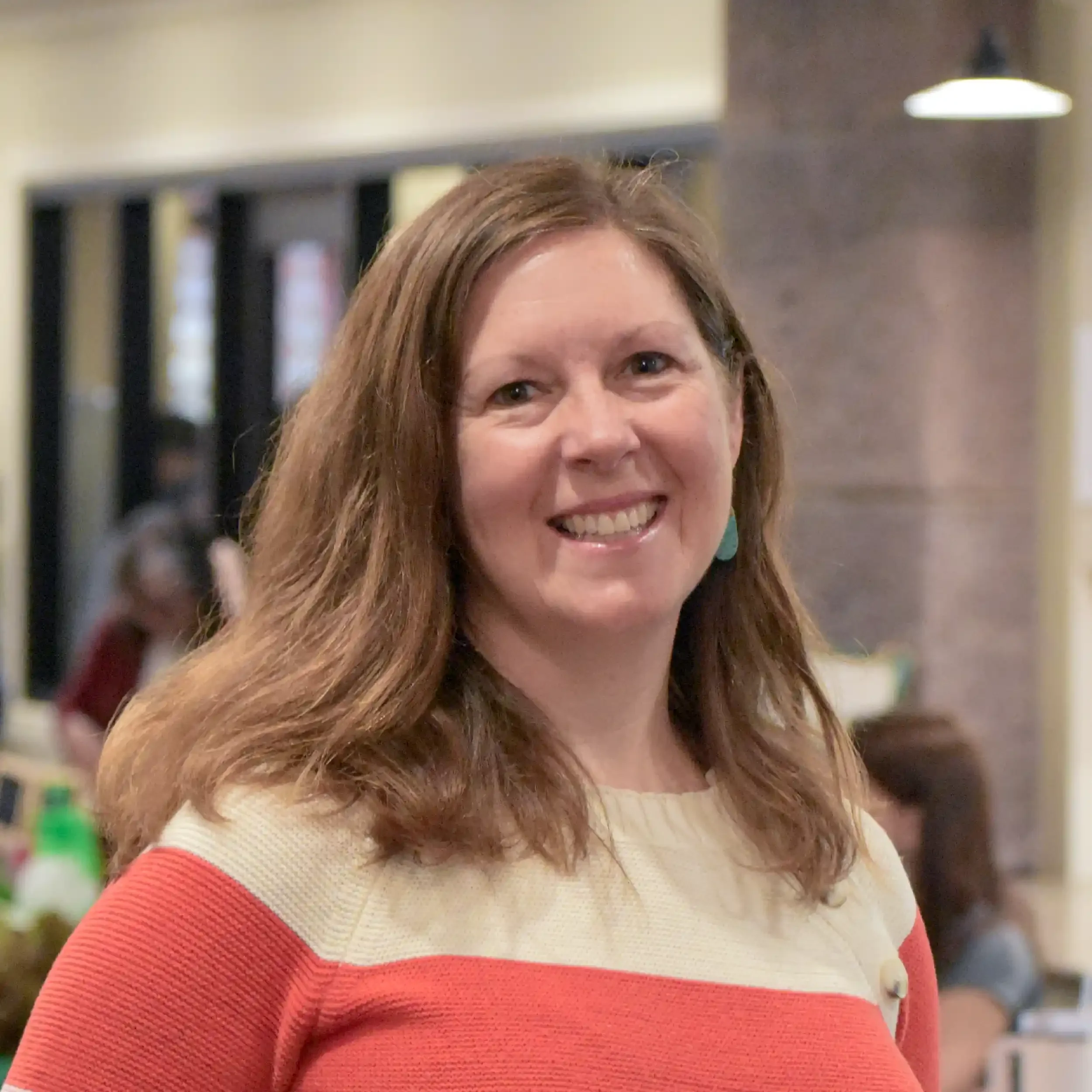 Close-up of a woman with long, curly brown hair smiling, wearing a cream and red striped sweater, in an indoor setting with blurred background.