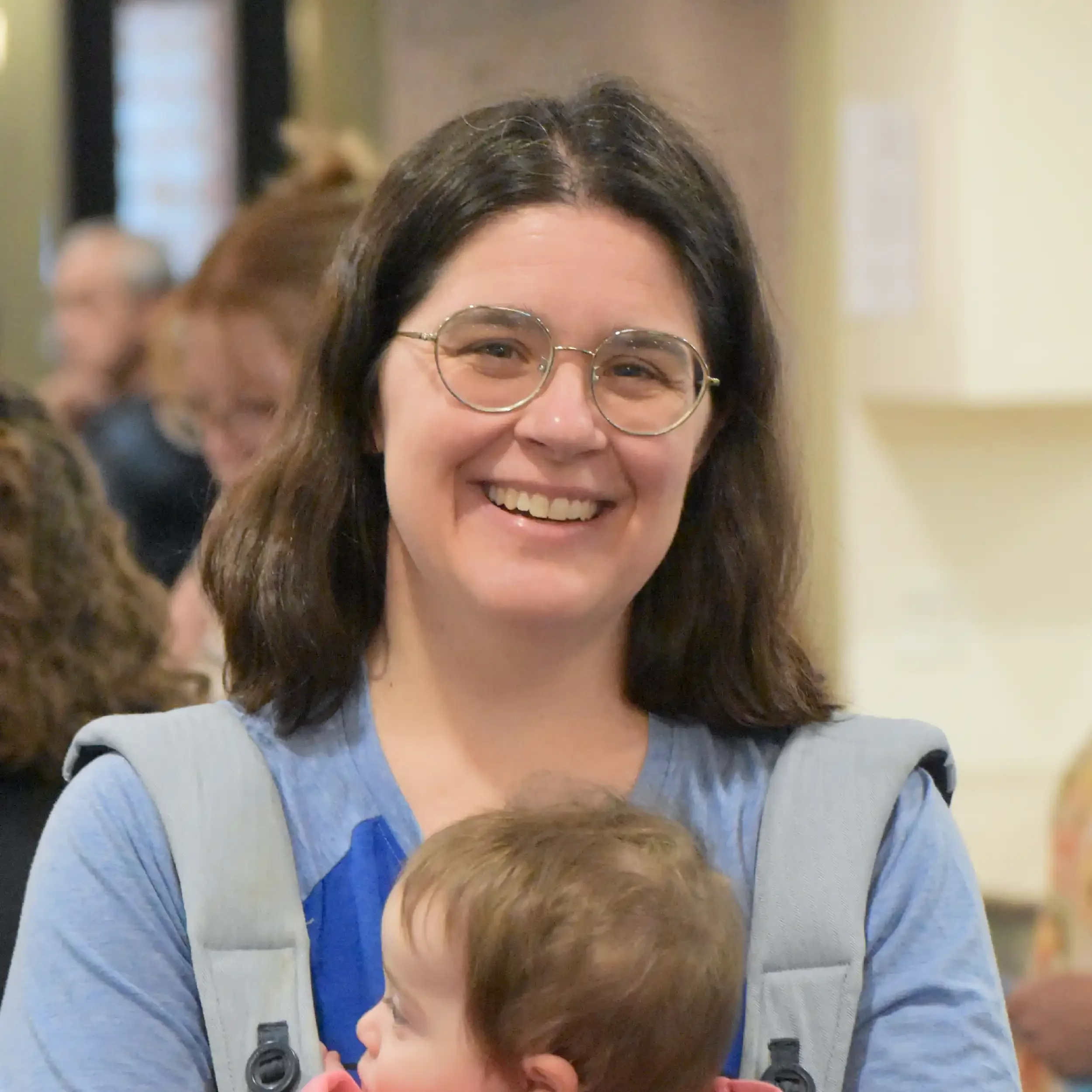 A woman with glasses and dark hair smiling, holding a young child with brown hair and a blue shirt, both indoors.