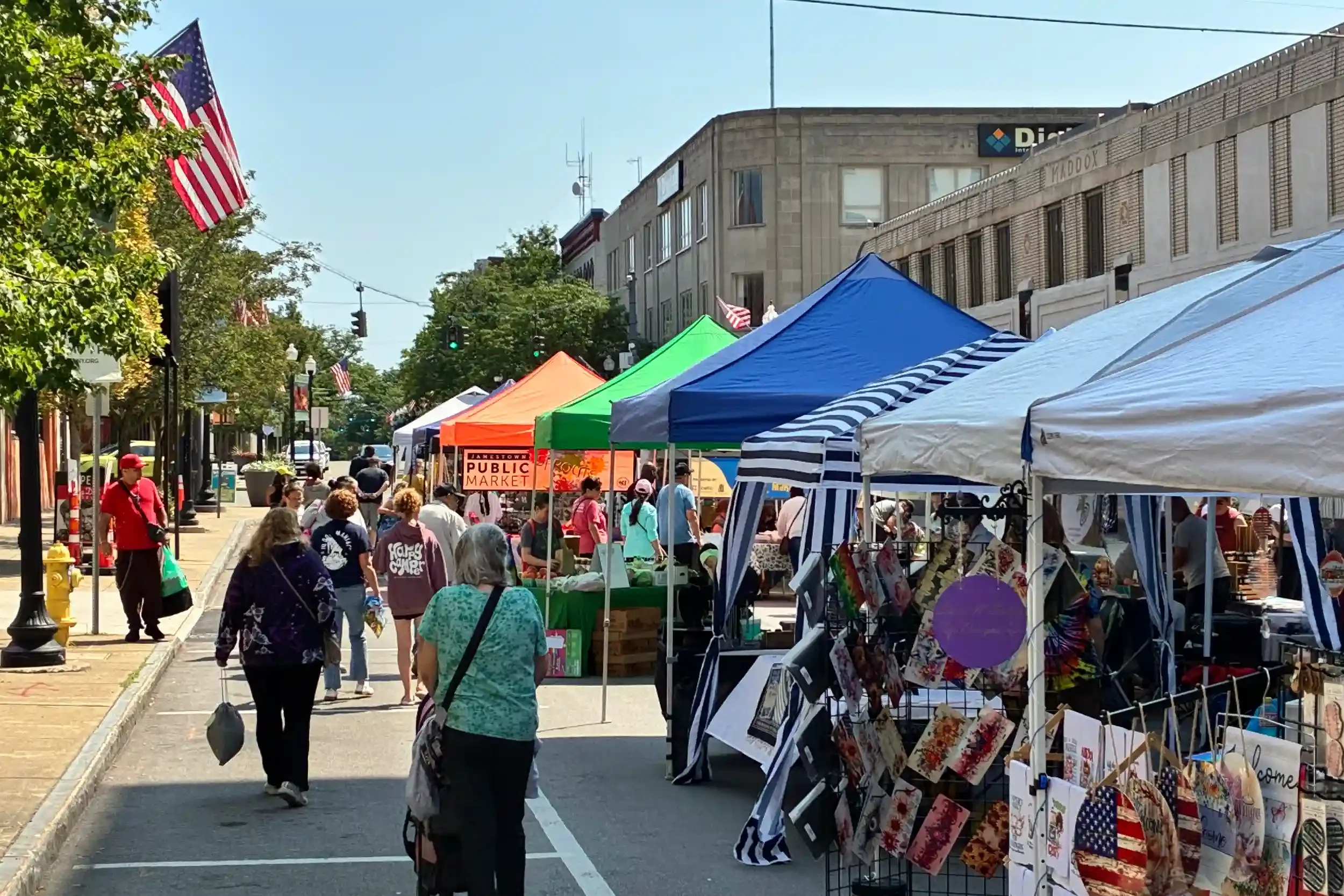 An outdoor street market with colorful tents, shoppers walking, and American flags displayed across the street buildings under a clear sky.