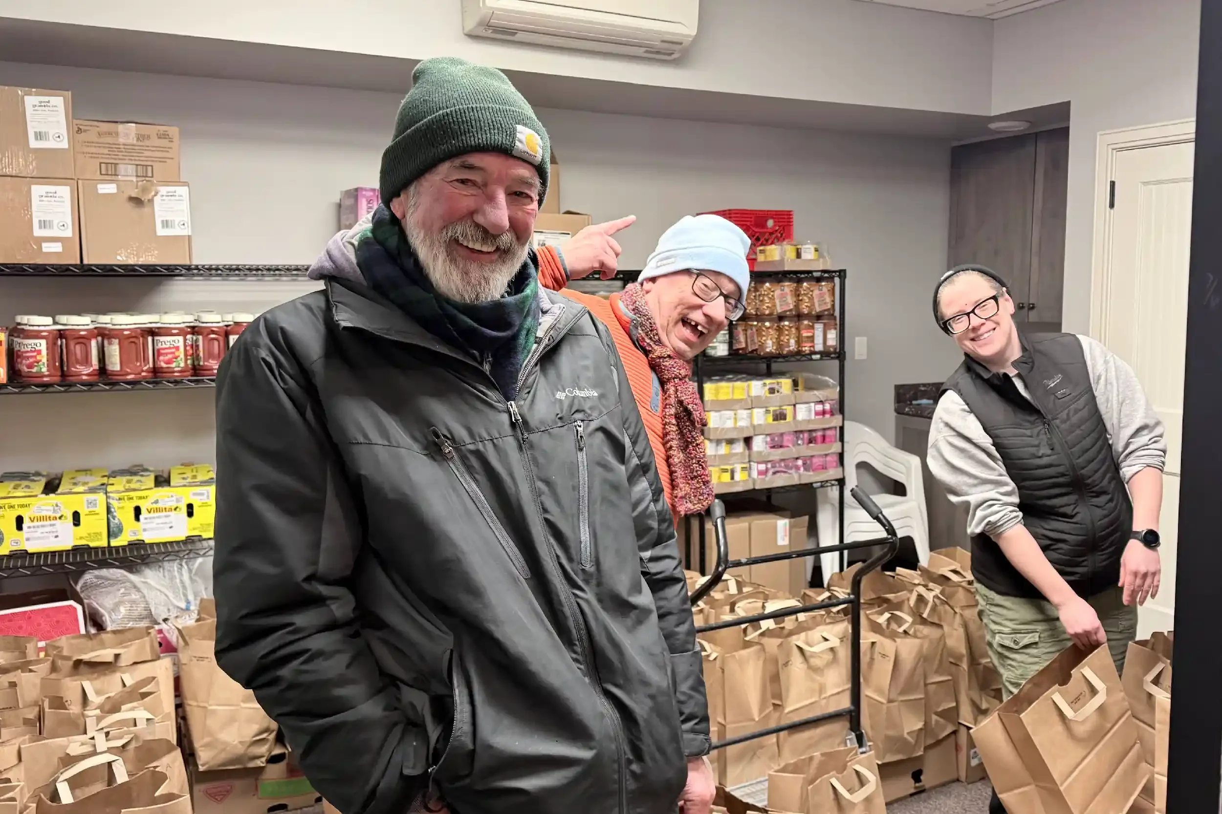 Three people smiling and posing in a grocery store or food pantry, surrounded by paper bags filled with food items on shelves and the floor.