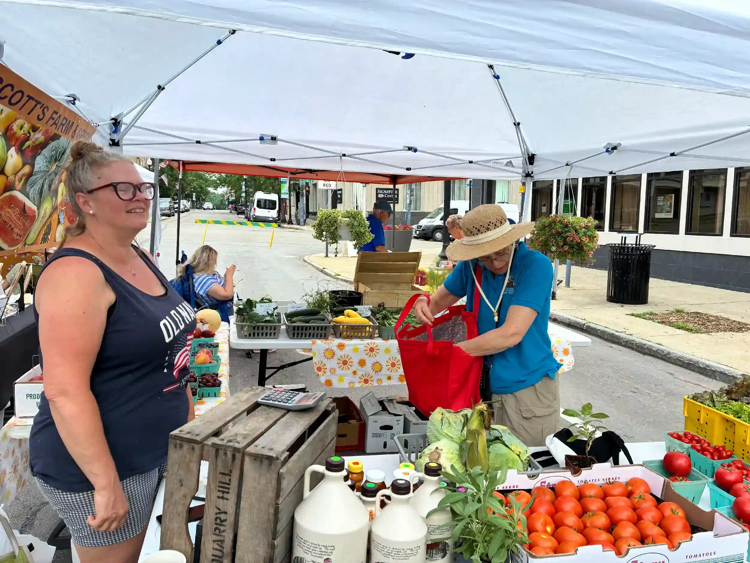 People shopping at an outdoor farmers market stall with fresh vegetables and produce, including tomatoes, lettuce, and herbs, under a white canopy on a city street.