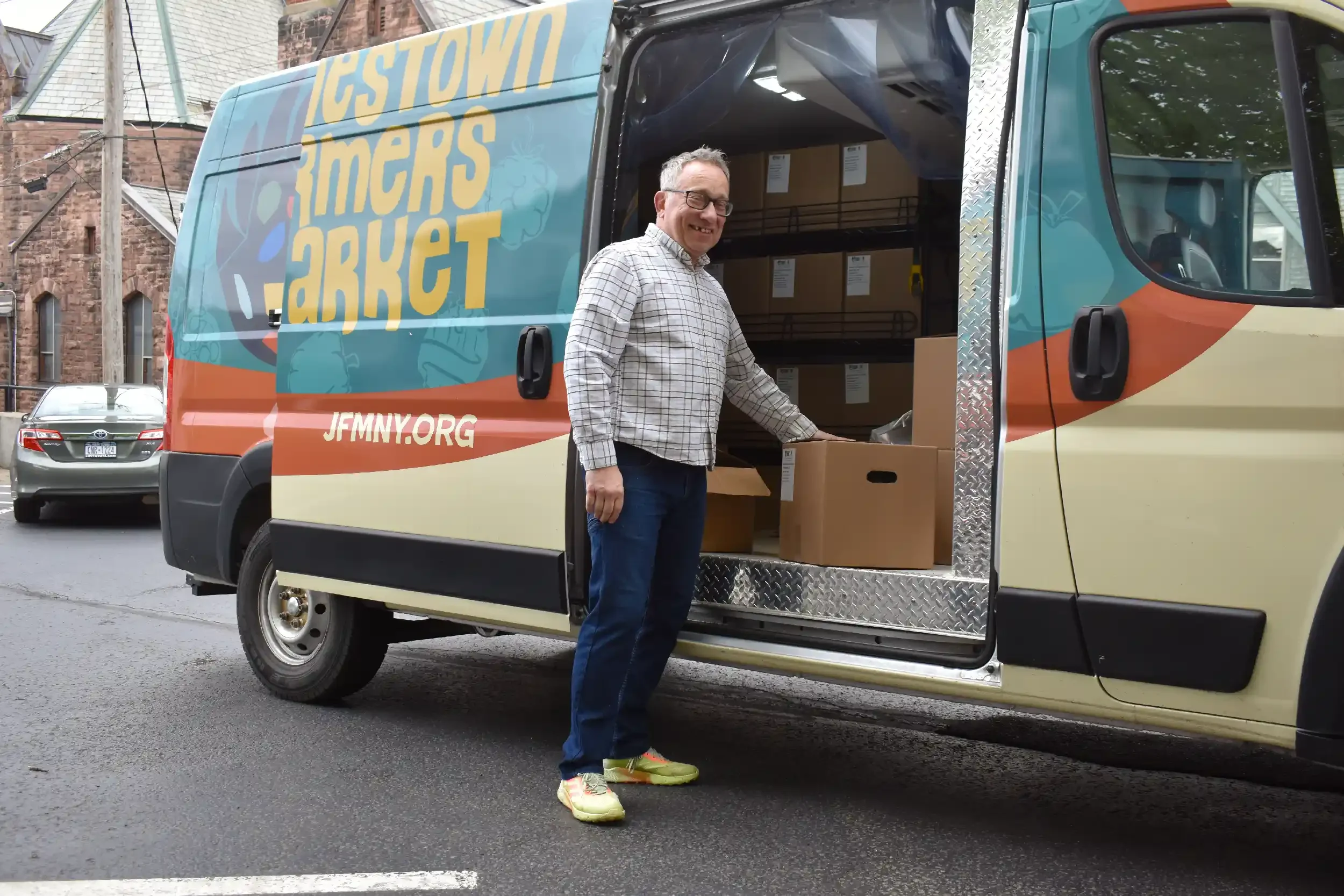 A man wearing glasses, a checkered shirt, and neon green shoes unloading cardboard boxes from a delivery van with a colorful design that reads "Tigstowli Farmers Market" and a website URL "JFMNY.ORG" in an urban neighborhood.