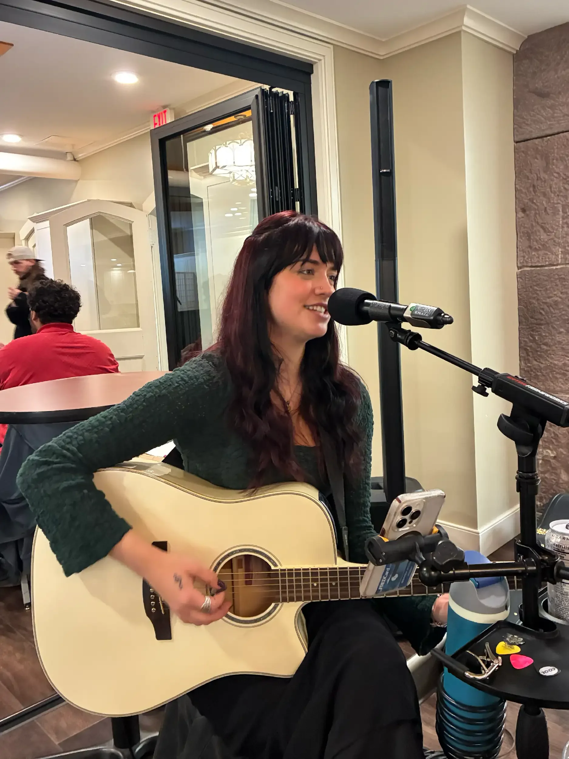 A woman with dark hair and a nose piercing is playing an acoustic guitar and singing into a microphone in an indoor setting. She is sitting at a table with a smartphone on it, and there are people in the background.