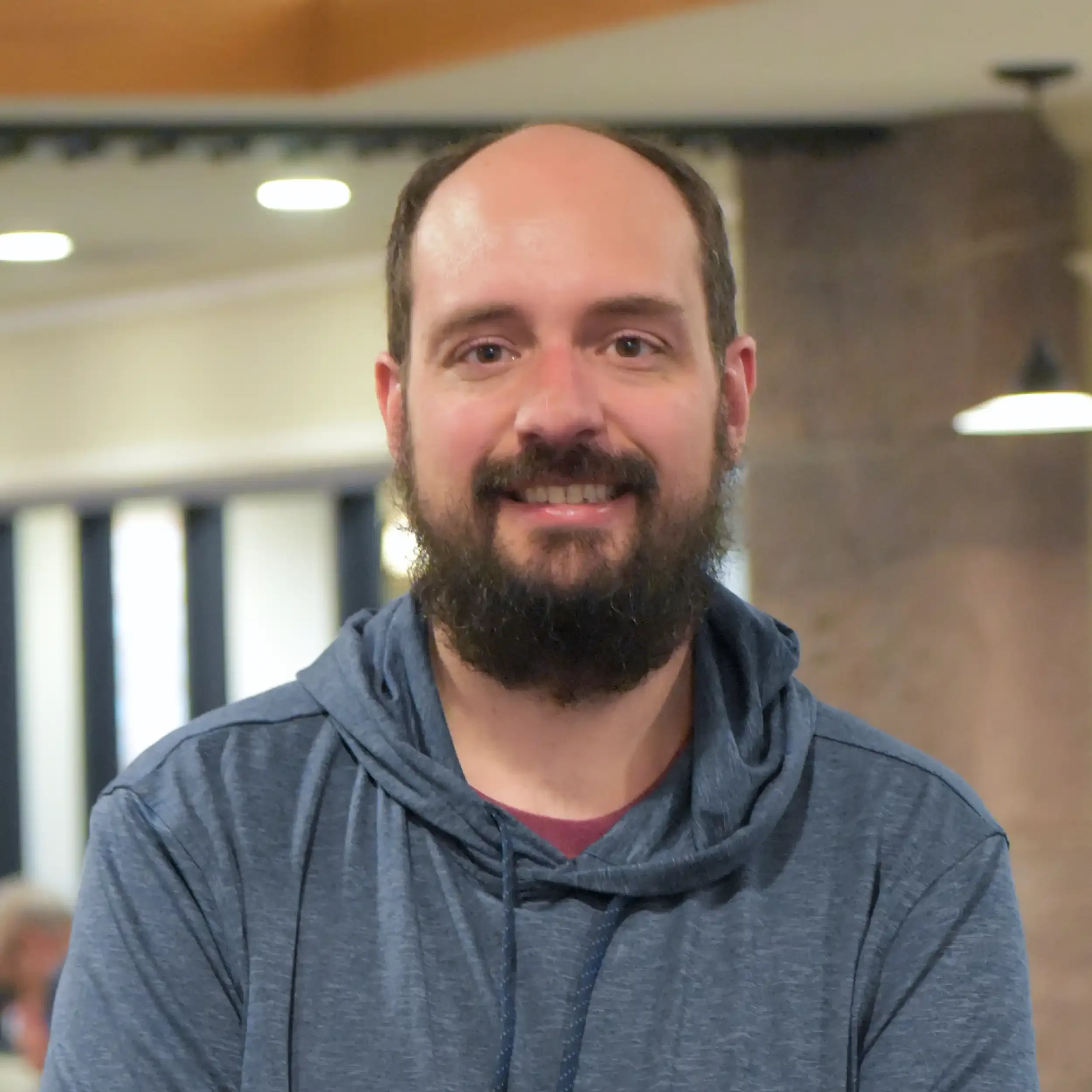 Close-up of a smiling man with a beard and mustache, wearing a gray jacket and black earbuds, indoors with ceiling lights and blurred background.