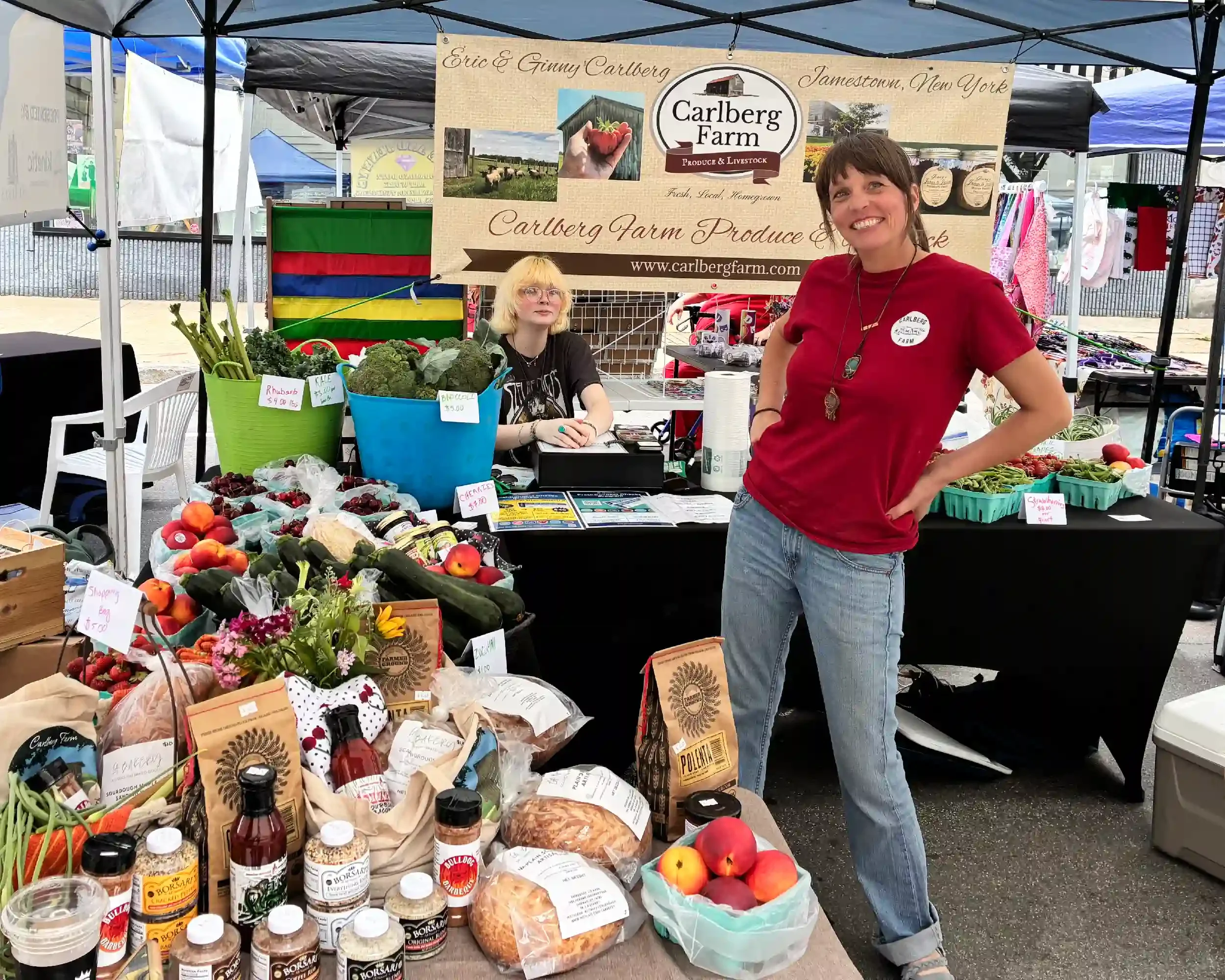 A woman in a red shirt standing next to a market stall with fresh produce at a farmers market. The stall has a large sign for Carlberg Farm, featuring pictures of farm scenes and the farm's website. The table has various vegetables like peaches, cherry tomatoes, broccoli, and herbs, along with bottles of sauces and other products. Another person with blonde hair and glasses is sitting behind the stall, with clothes hanging in the background.