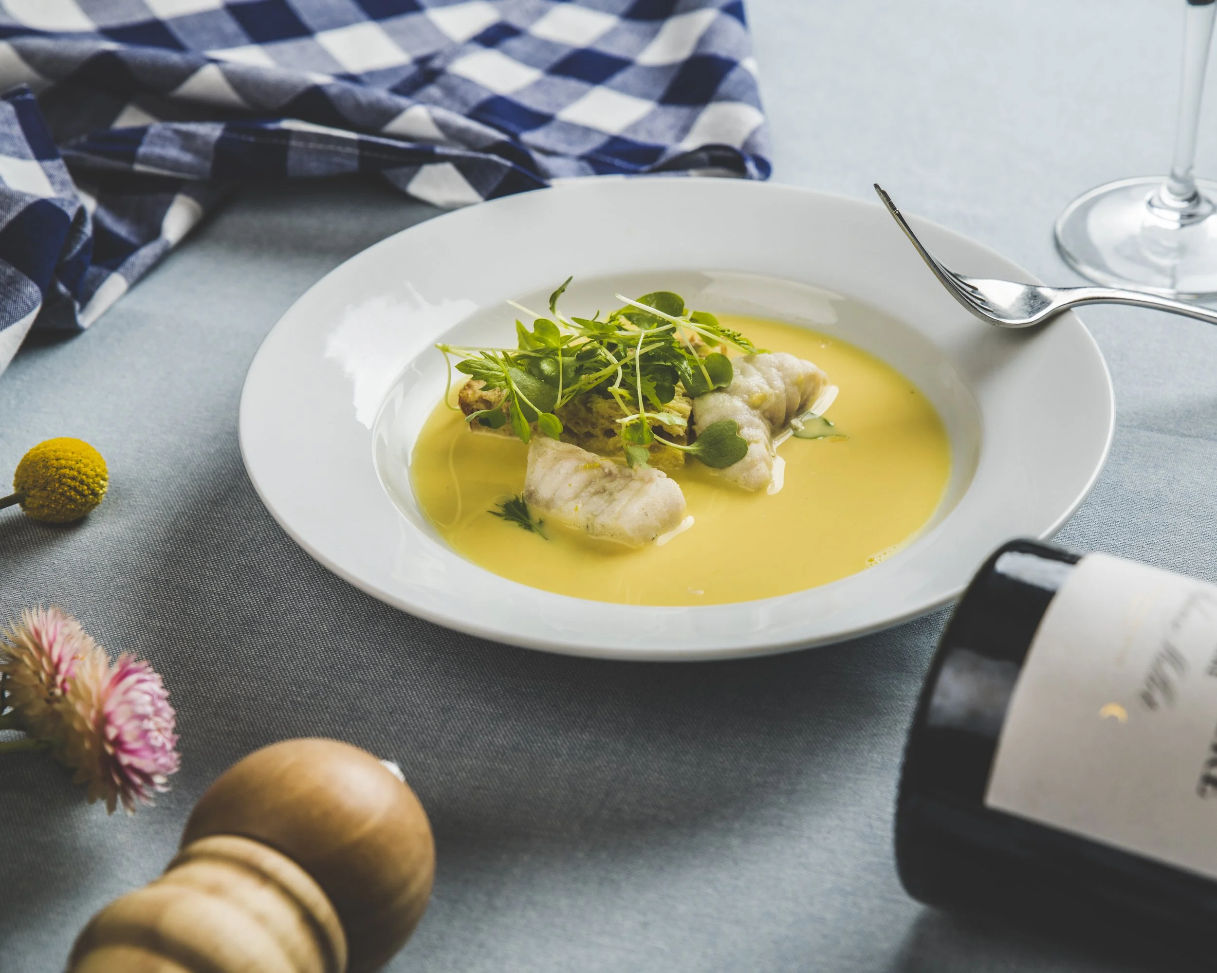 A plate of cooked fish garnished with greens and served with a yellow sauce on a table with a checkered cloth, a fork, a glass of wine, and flowers nearby.