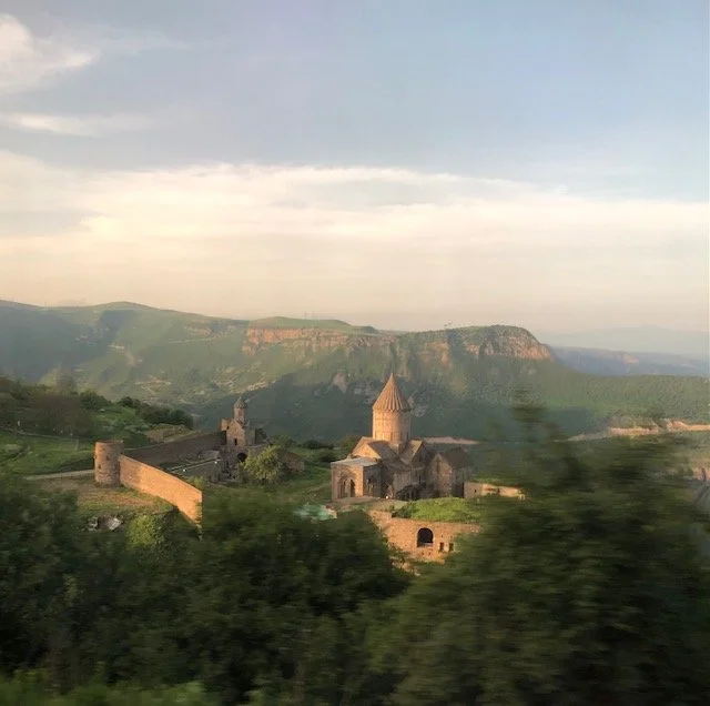 A scenic view of an ancient stone monastery and surrounding fortress walls on a green hill, with mountains in the background under a partly cloudy sky.
