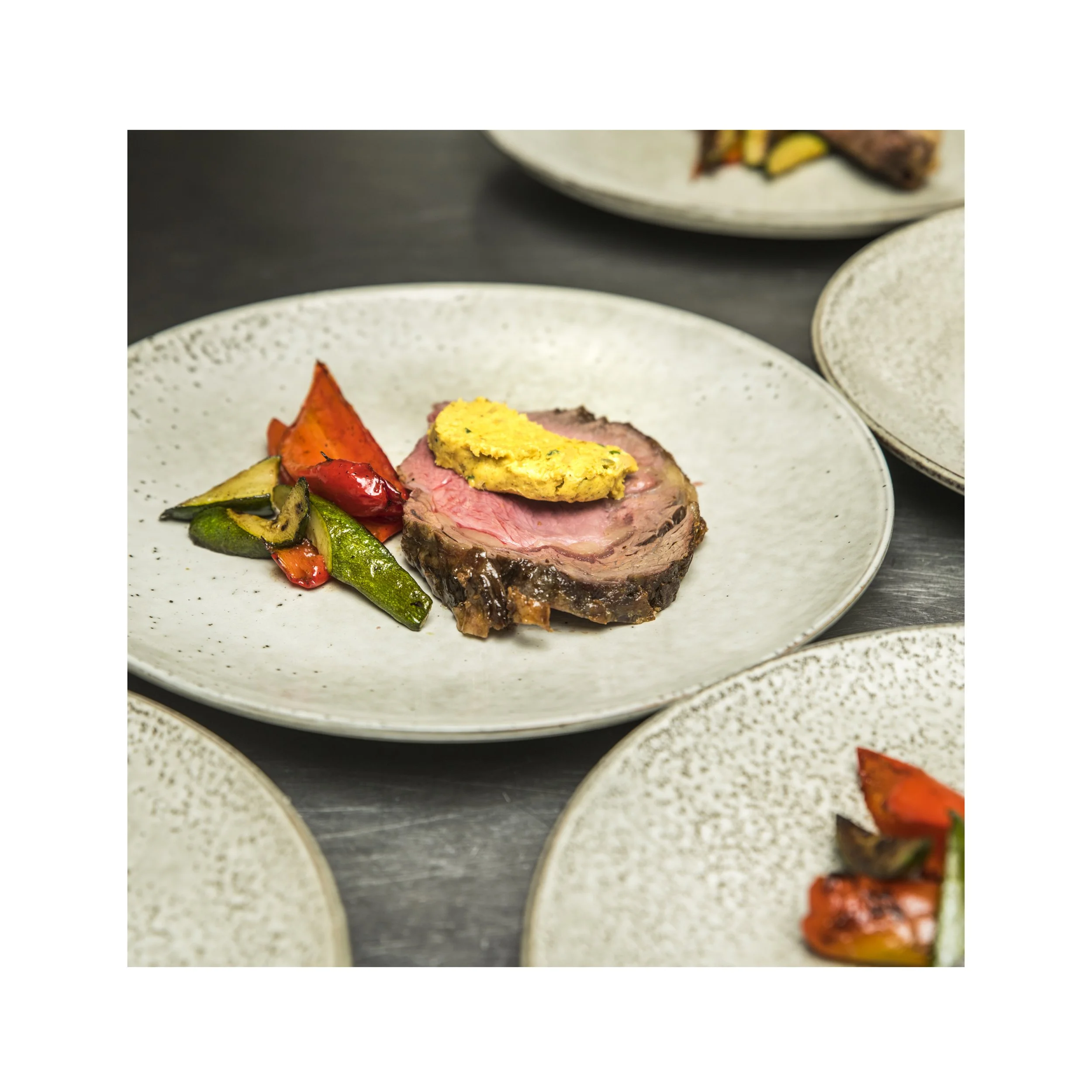 Close-up of a white speckled plate with roasted vegetables and pieces of cooked meat topped with cafe de Paris butter, with parts of similar plates visible in the background.