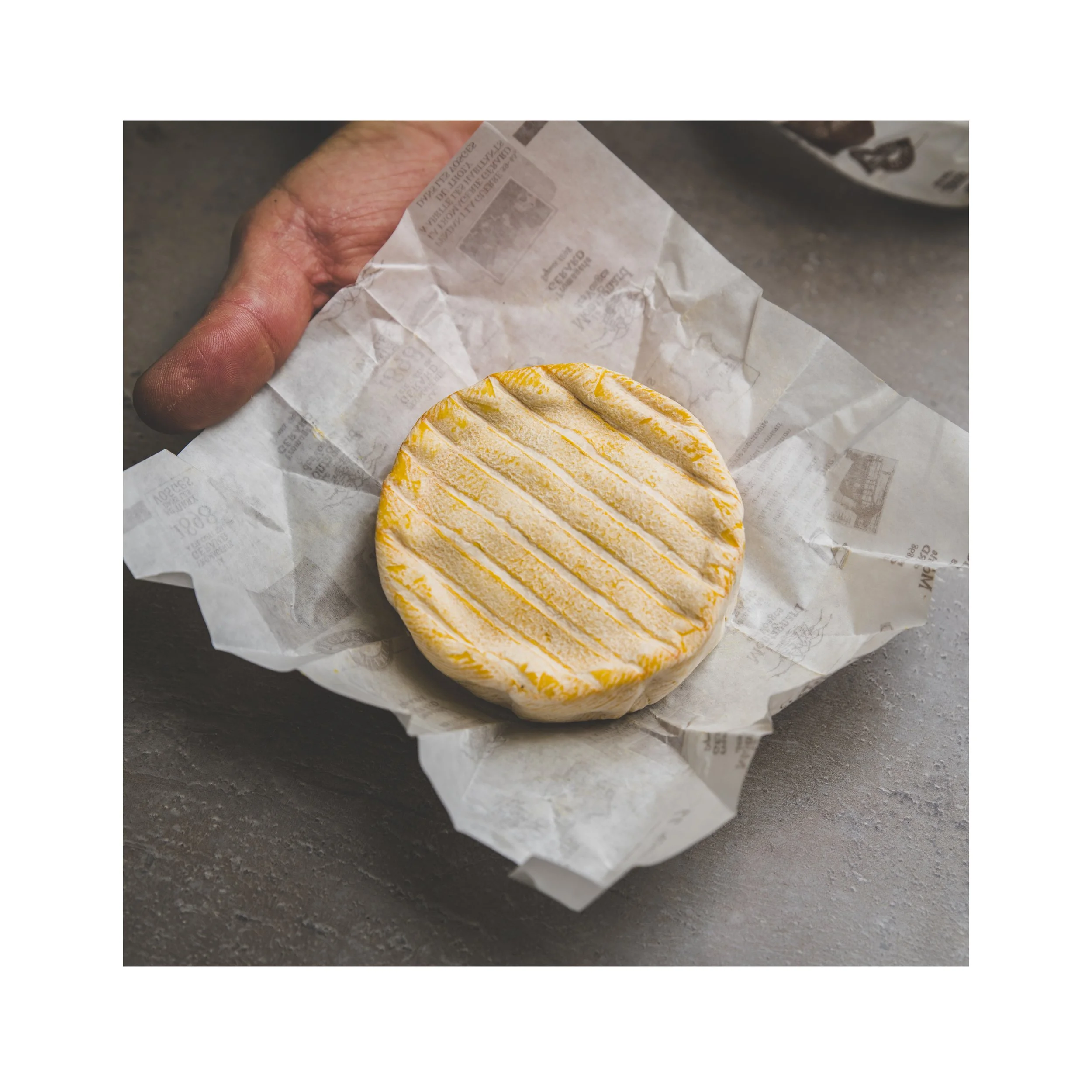 Close-up of a hand holding crinkled paper with a yellow and white striped fruit or cheese on top, on a gray surface with partial view of a patterned plate in the background.