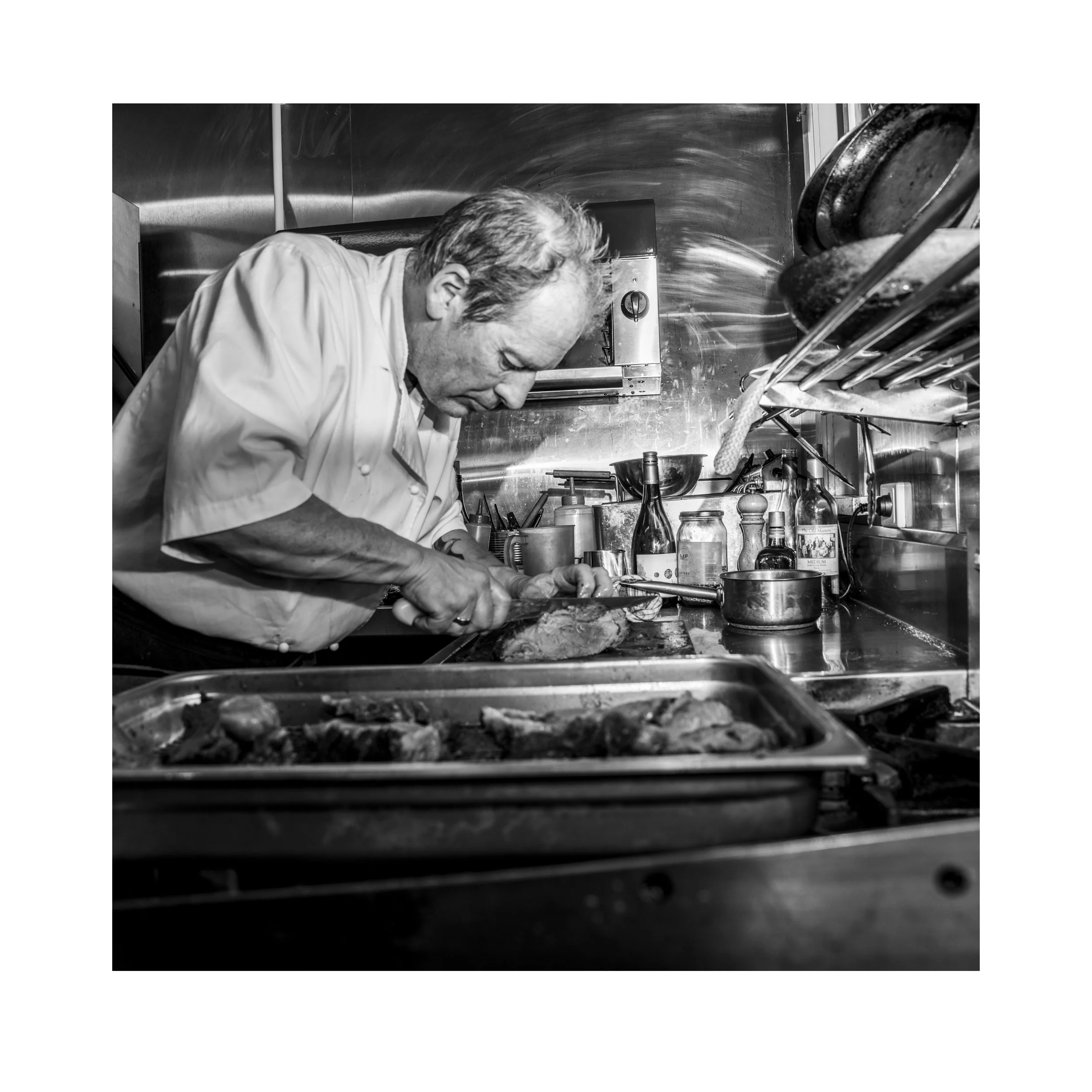 Chef Alain Hauswirth preparing food in a professional kitchen, with various bottles and kitchen utensils on the counter.
