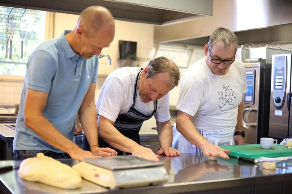 Three men working together in a professional kitchen, kneading dough on a counter.