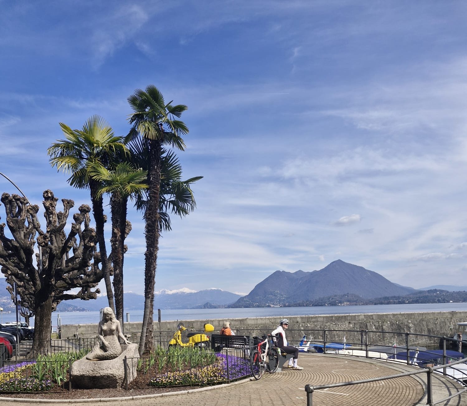 A lakeside scene with a statue of a mermaid, palm trees, and a mountain in the background. There are benches, a few people, bikes, and boats along the waterfront.