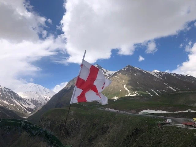 A mountain landscape with snow-capped peaks and a cloudy sky. A flag with a red cross on a white background is flying in the foreground.