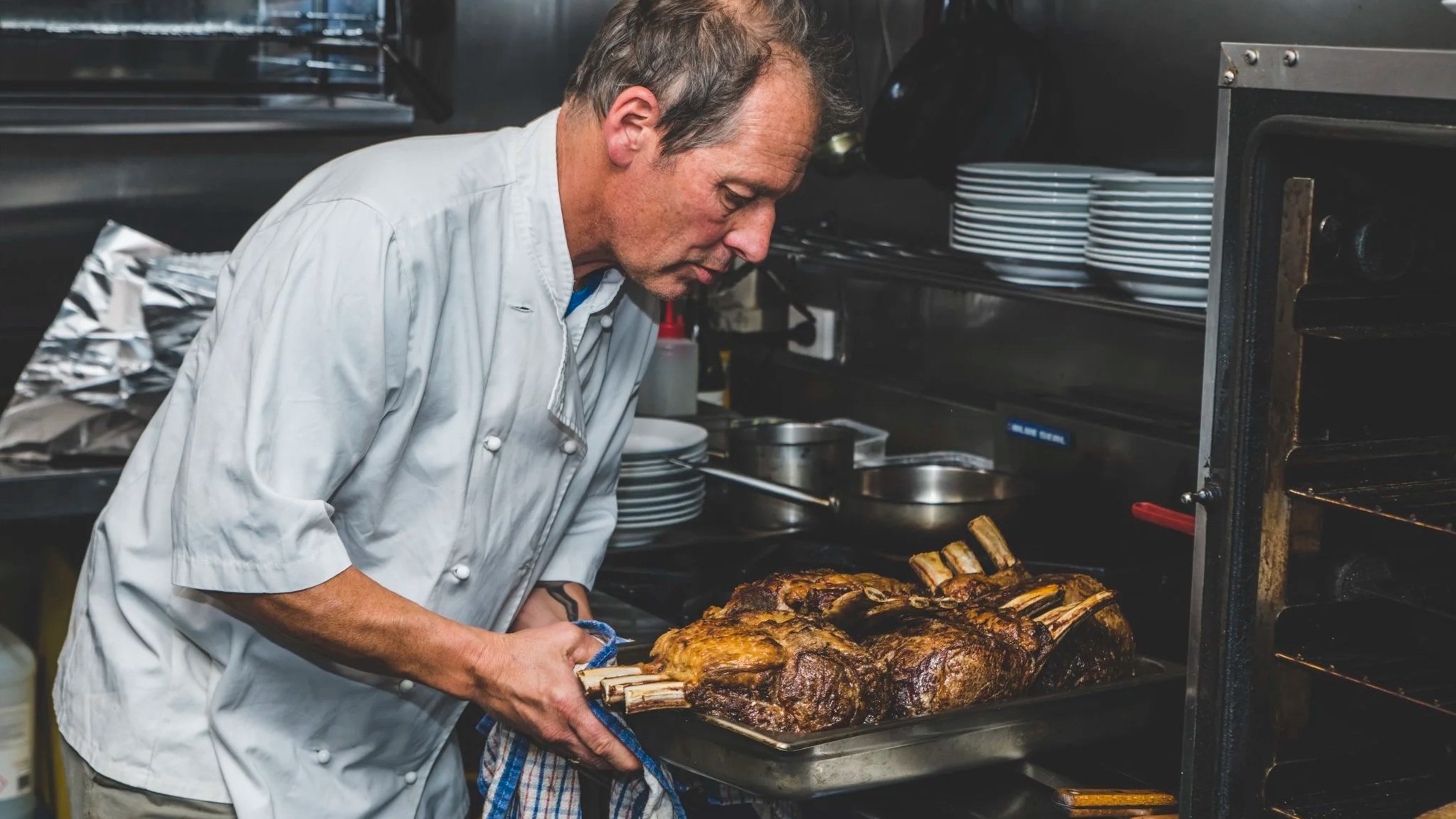 Chef Alain Hauswirth, in a white chef's coat taking a roasted piece of meat out of an oven in a professional kitchen. The oven door is open, and the kitchen has stainless steel shelves with plates and cooking utensils.