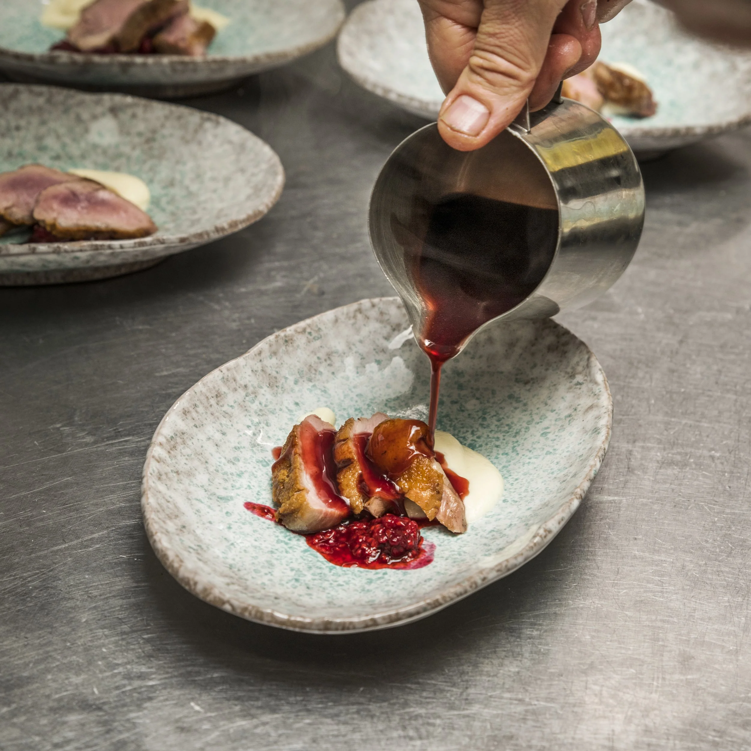 A person pouring red sauce onto seared duck, on decorative plates.