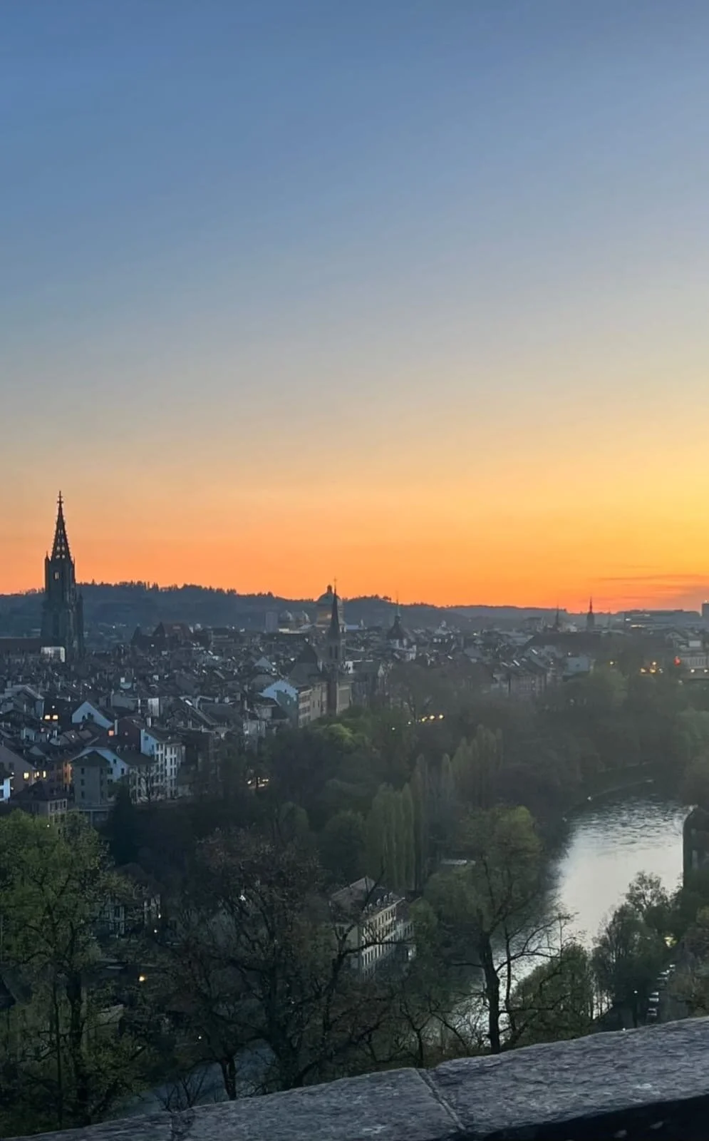 A cityscape at sunset featuring a river, rooftops, and church steeples, with a colorful sky transitioning from blue to orange.